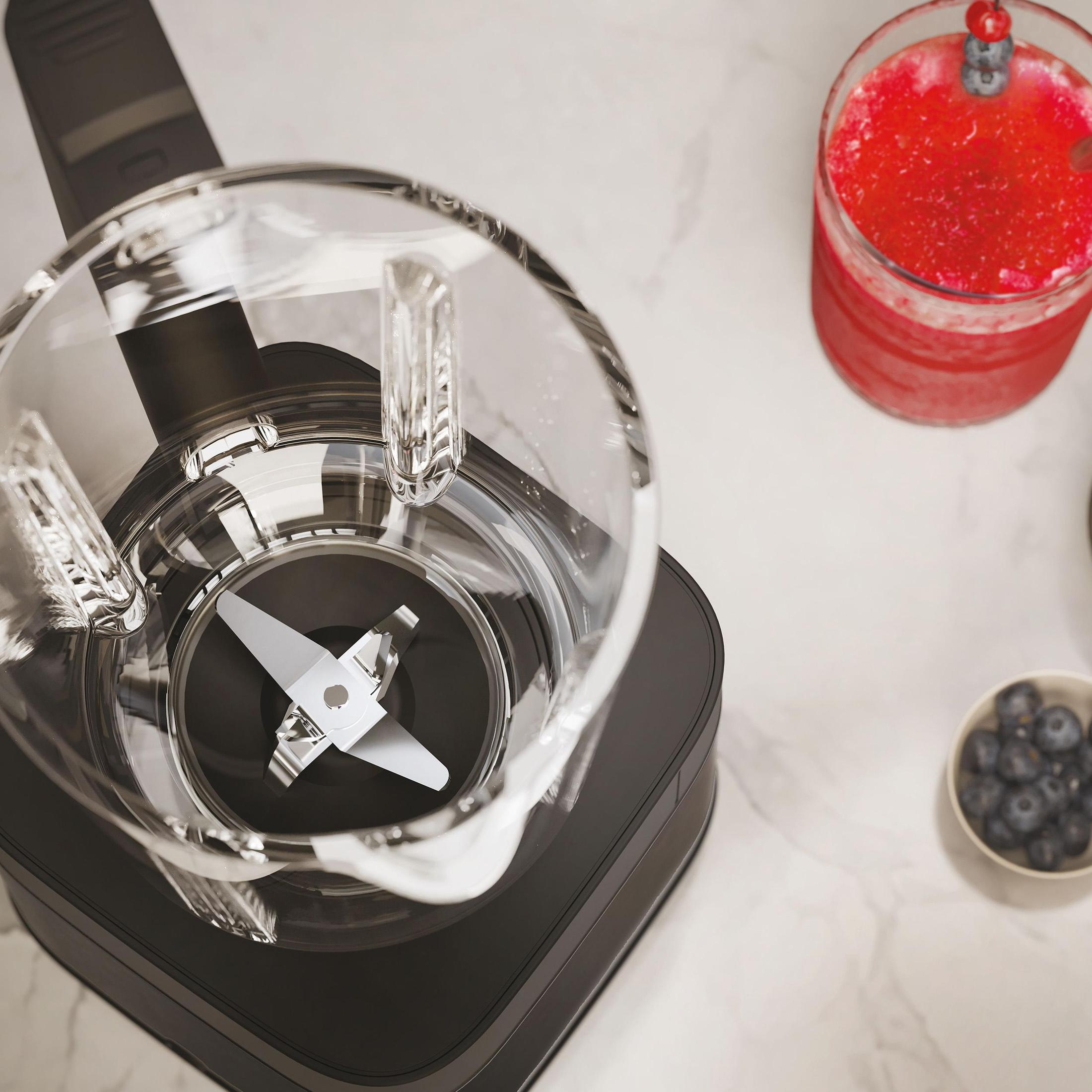 Blender z przezroczystym dzbankiem z ostrzem. Czerwony napój w szklance z jagodami i miska jagód.