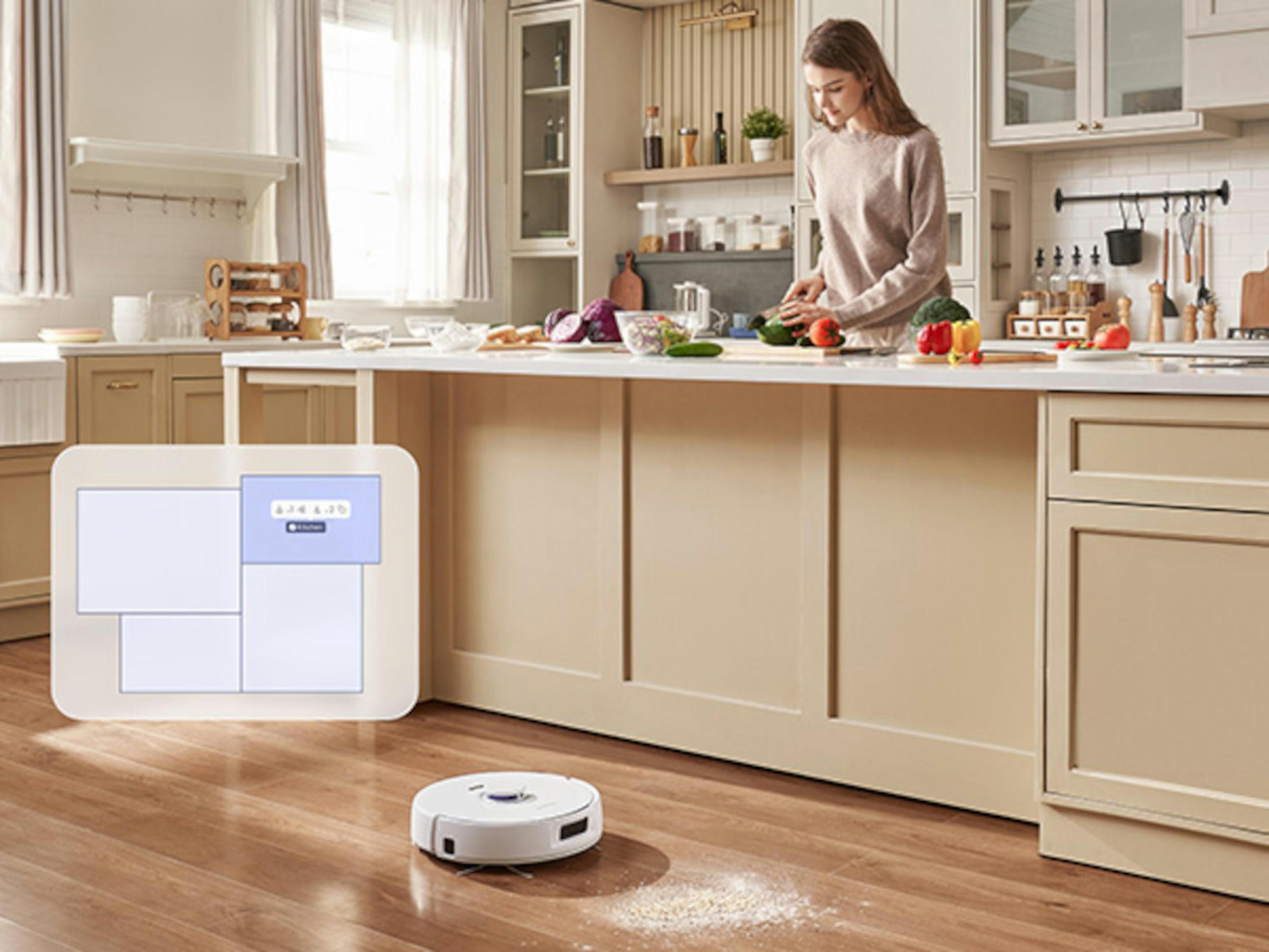 Donna in cucina, taglia le verdure. Un robot aspirapolvere e un'isola cucina sono in vista.