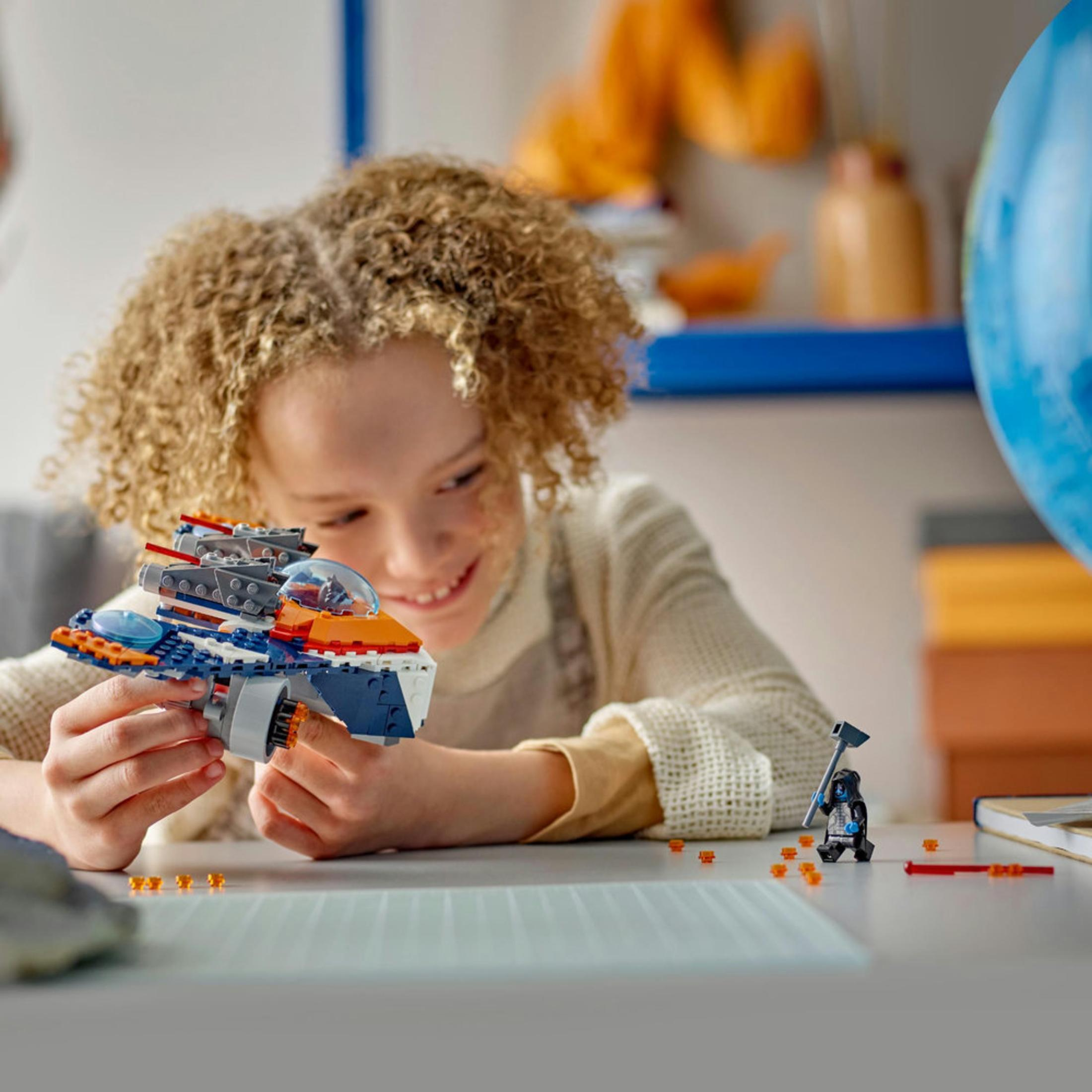 Genç bir kız, bir Lego uçağıyla oynarken gülümsüyor. Bazı lego figürleri ve bir küre görülebilir.