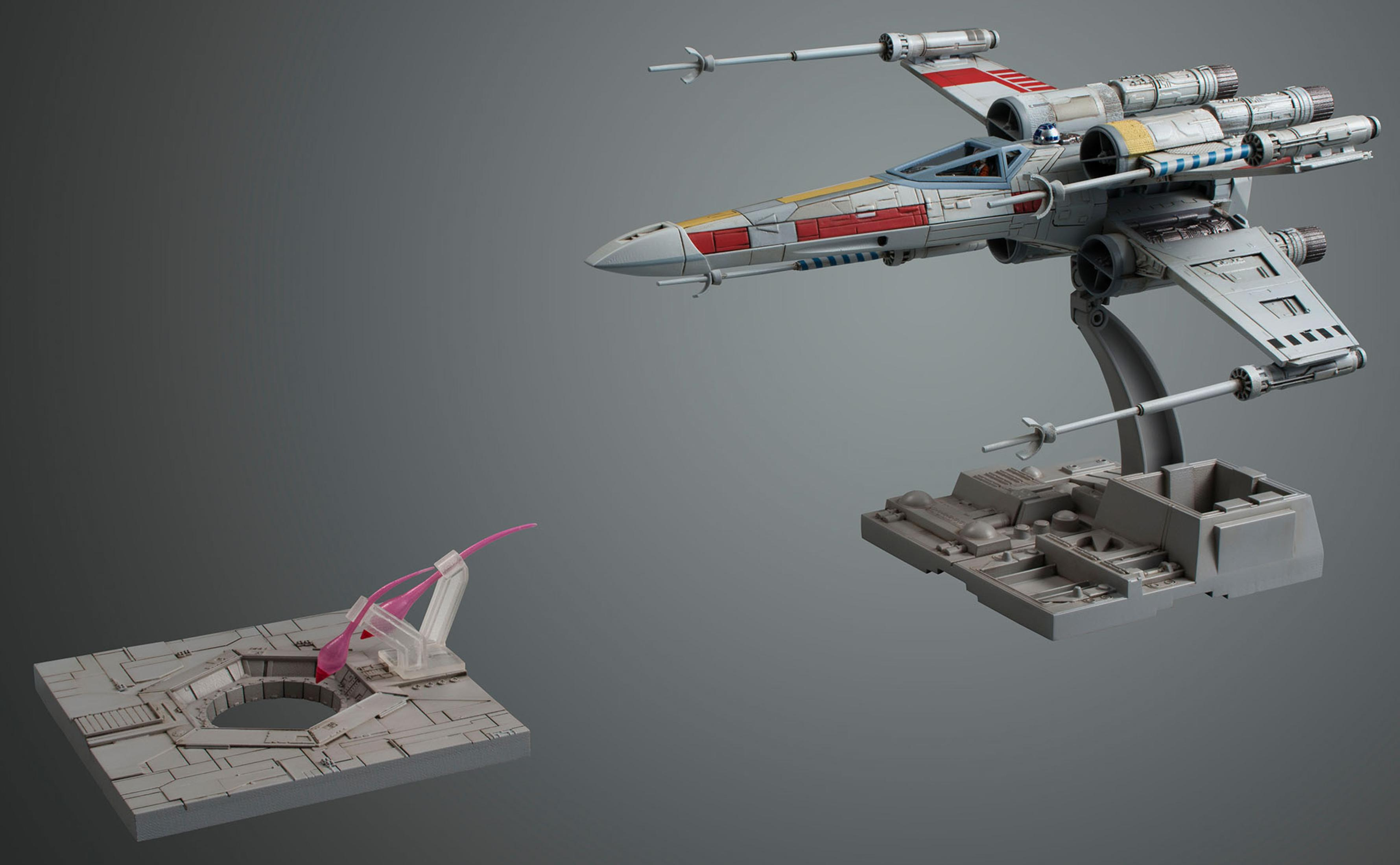 Modello di caccia X-wing su supporto, due basi modello, una con supporti rosa, sfondo grigio.