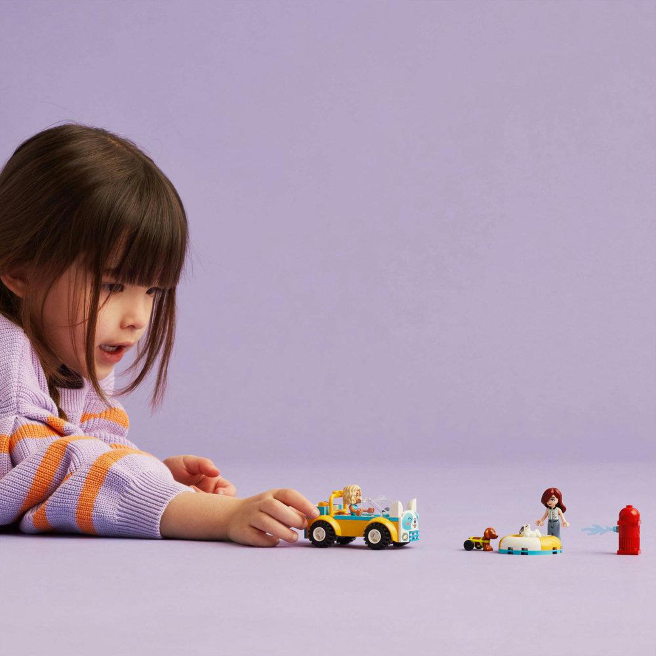 Dziecko patrzy na scenę Lego z żółtym samochodem, figurkami i hydrantem na fioletowym tle.