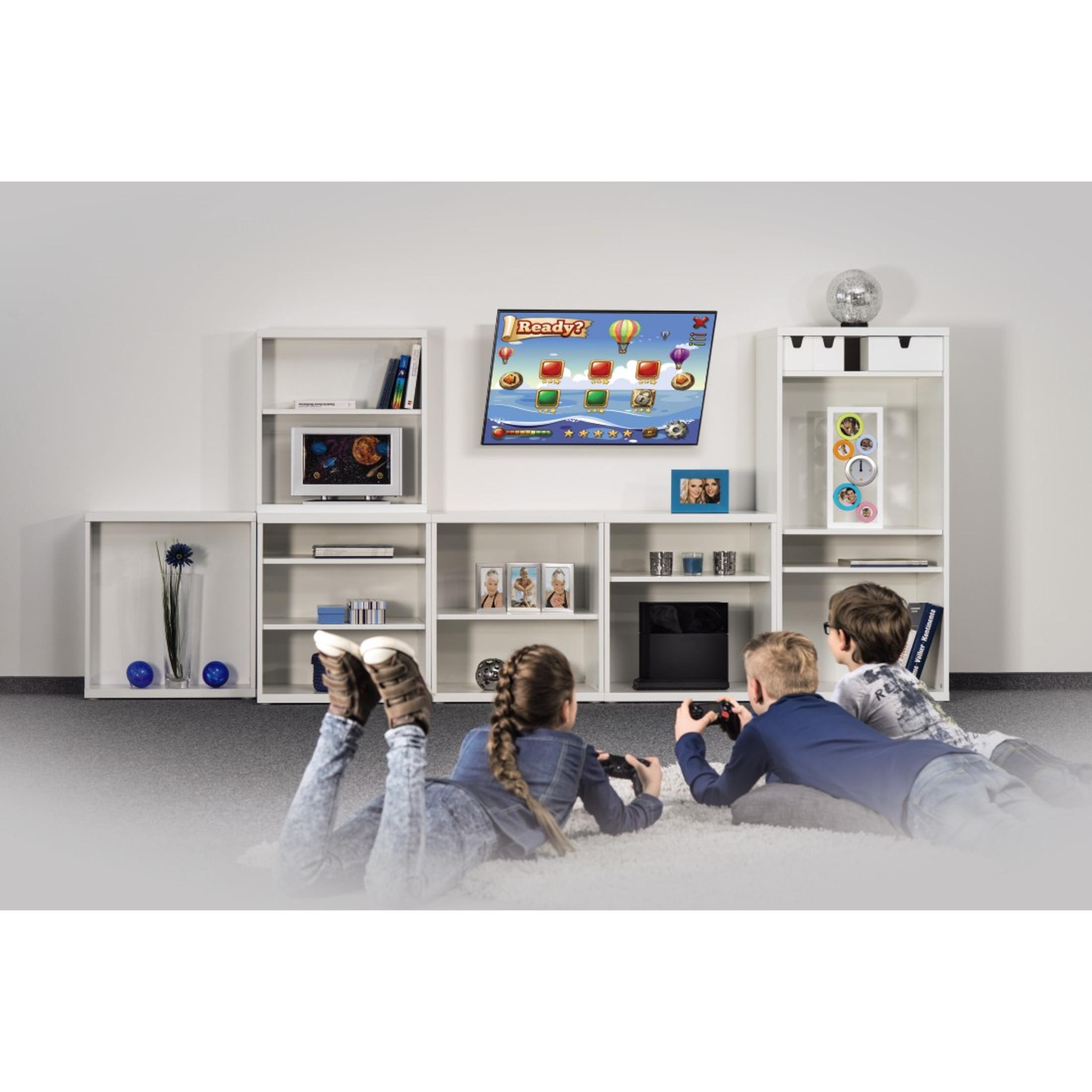 Niños jugando videojuegos en una sala de estar. Un televisor está montado en la pared. Estanterías blancas a los lados.