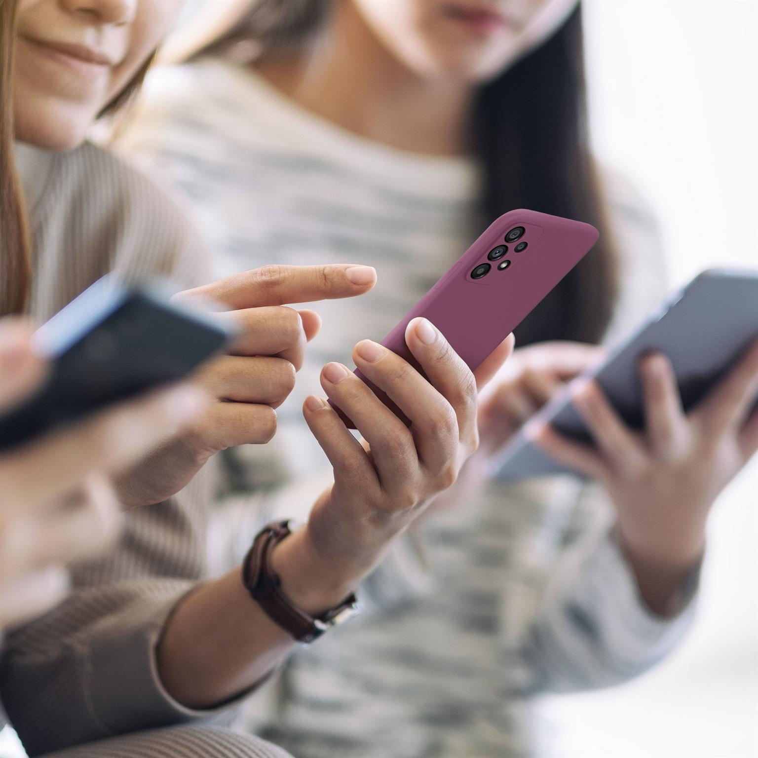 Drei Personen halten Smartphones. Ein Telefon ist lila, andere schwarz und grau. Verschwommener Hintergrund.