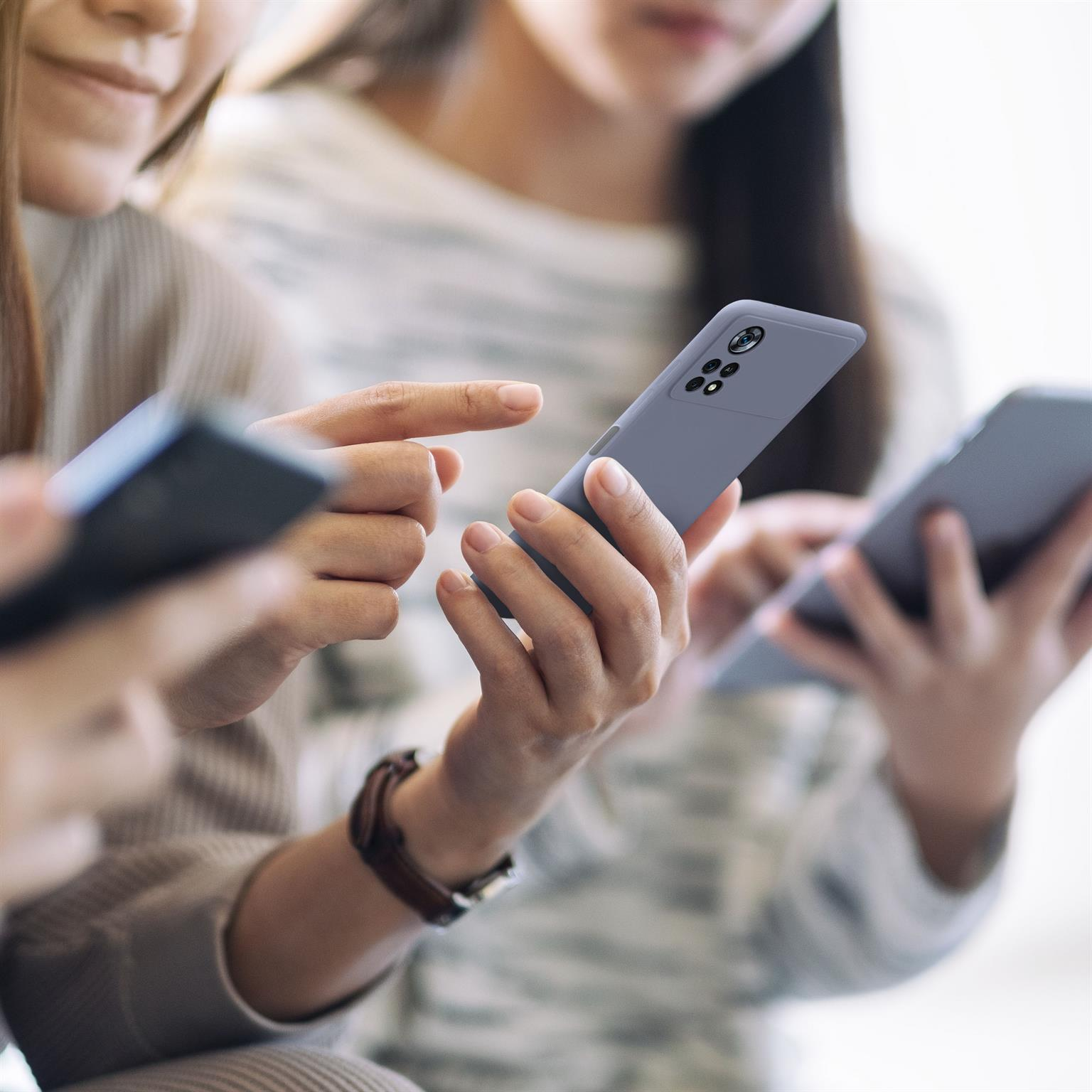 Drei Personen halten Telefone, eine zeigt. Telefone sind grau. Hintergrund ist unscharf.