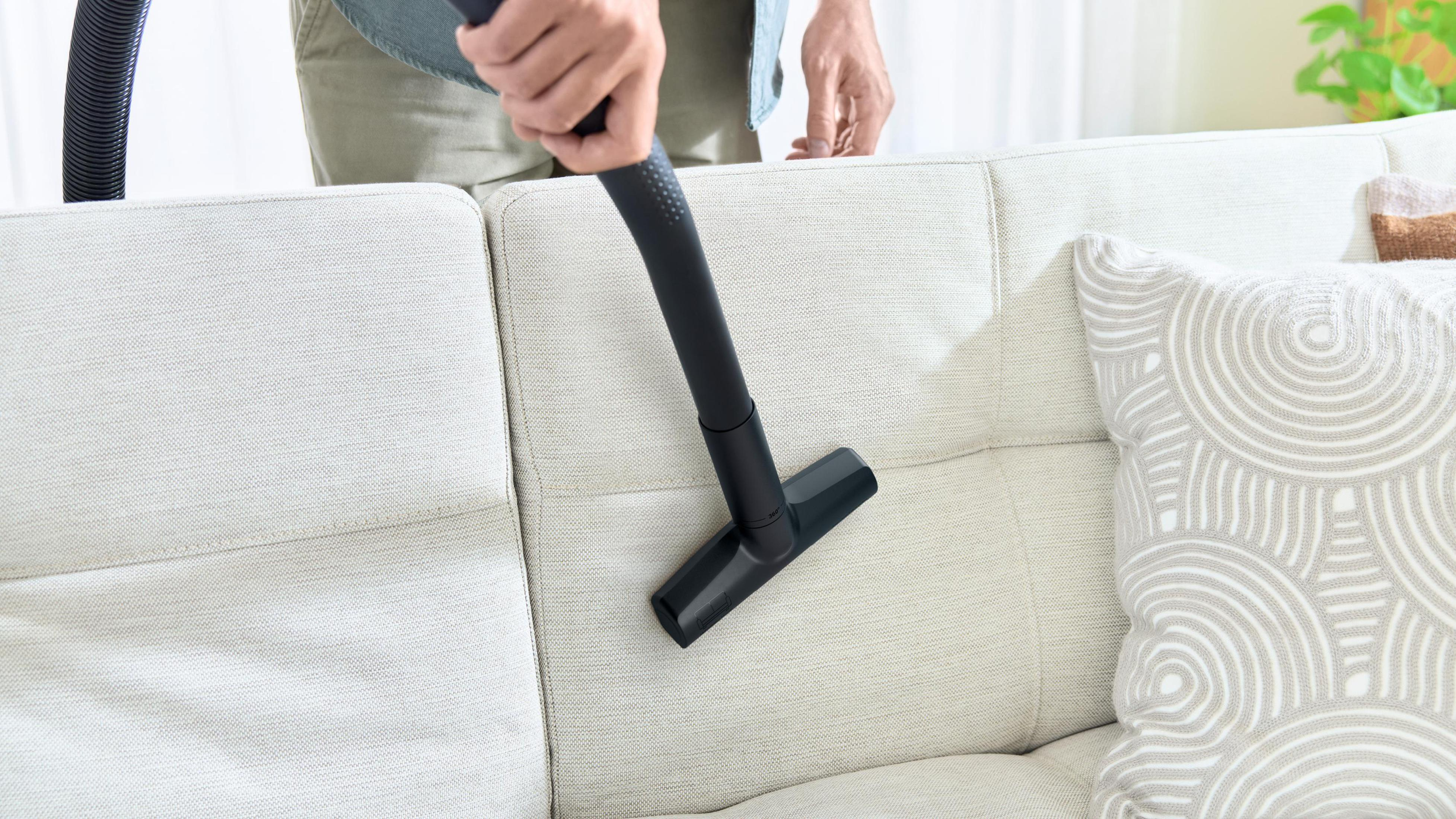 Osoba odkurza białą sofę czarną głowicą odkurzacza.