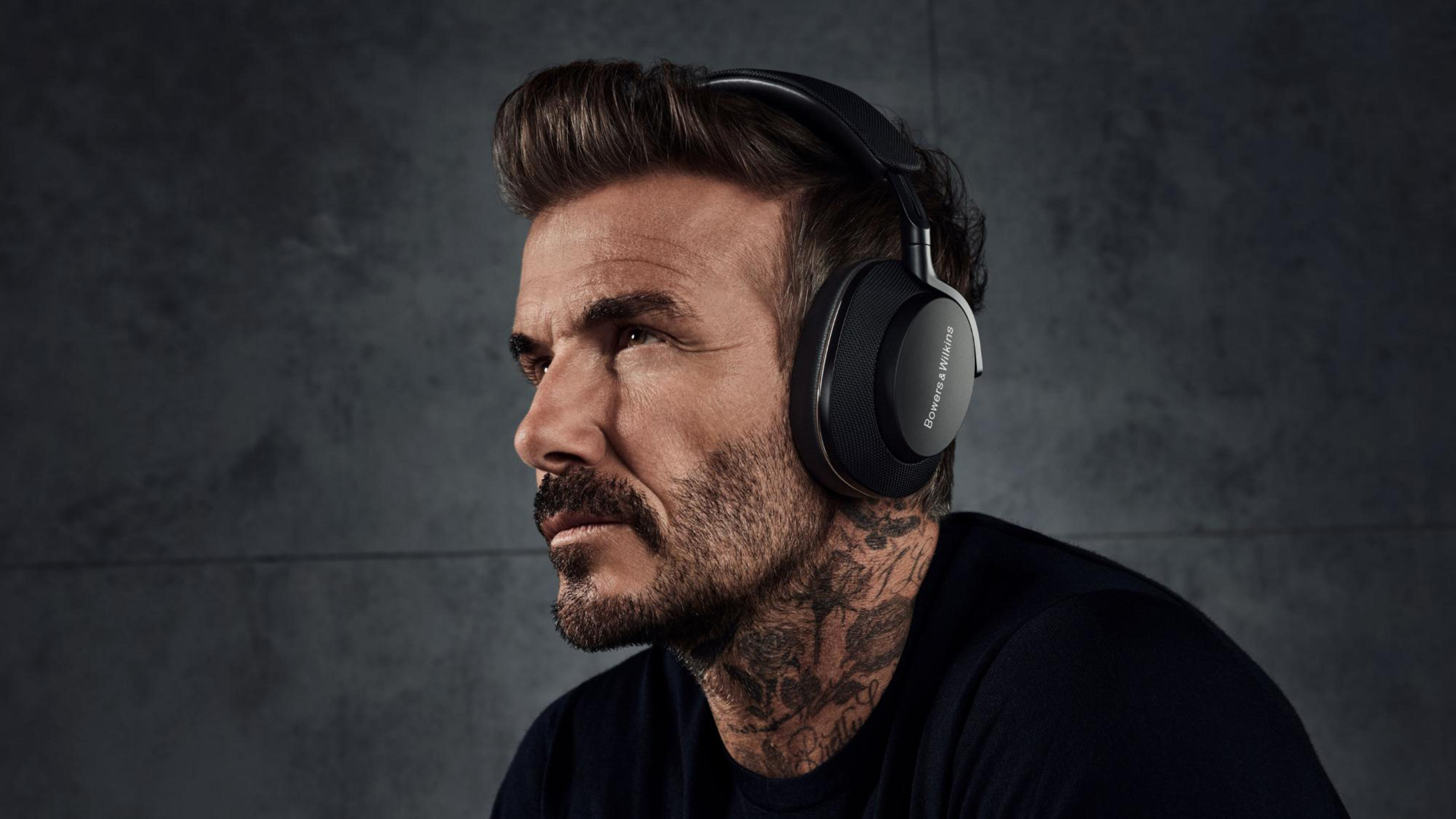 David Beckham w czarnych słuchawkach, czarnej koszuli, z tatuażami na szyi i szarym tle.