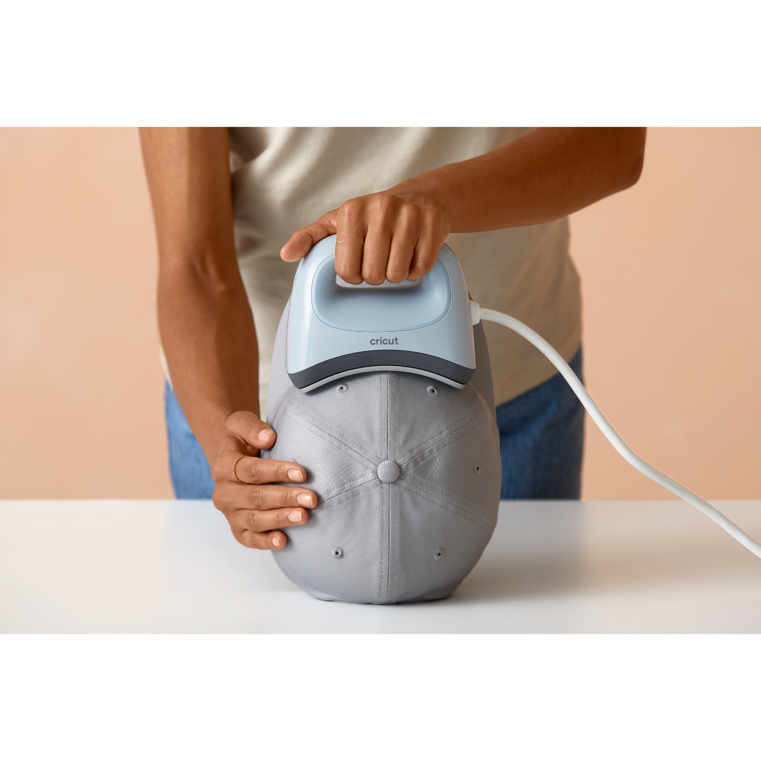 Una persona usa una pressa a caldo Cricut su un berretto da baseball grigio su una superficie bianca.