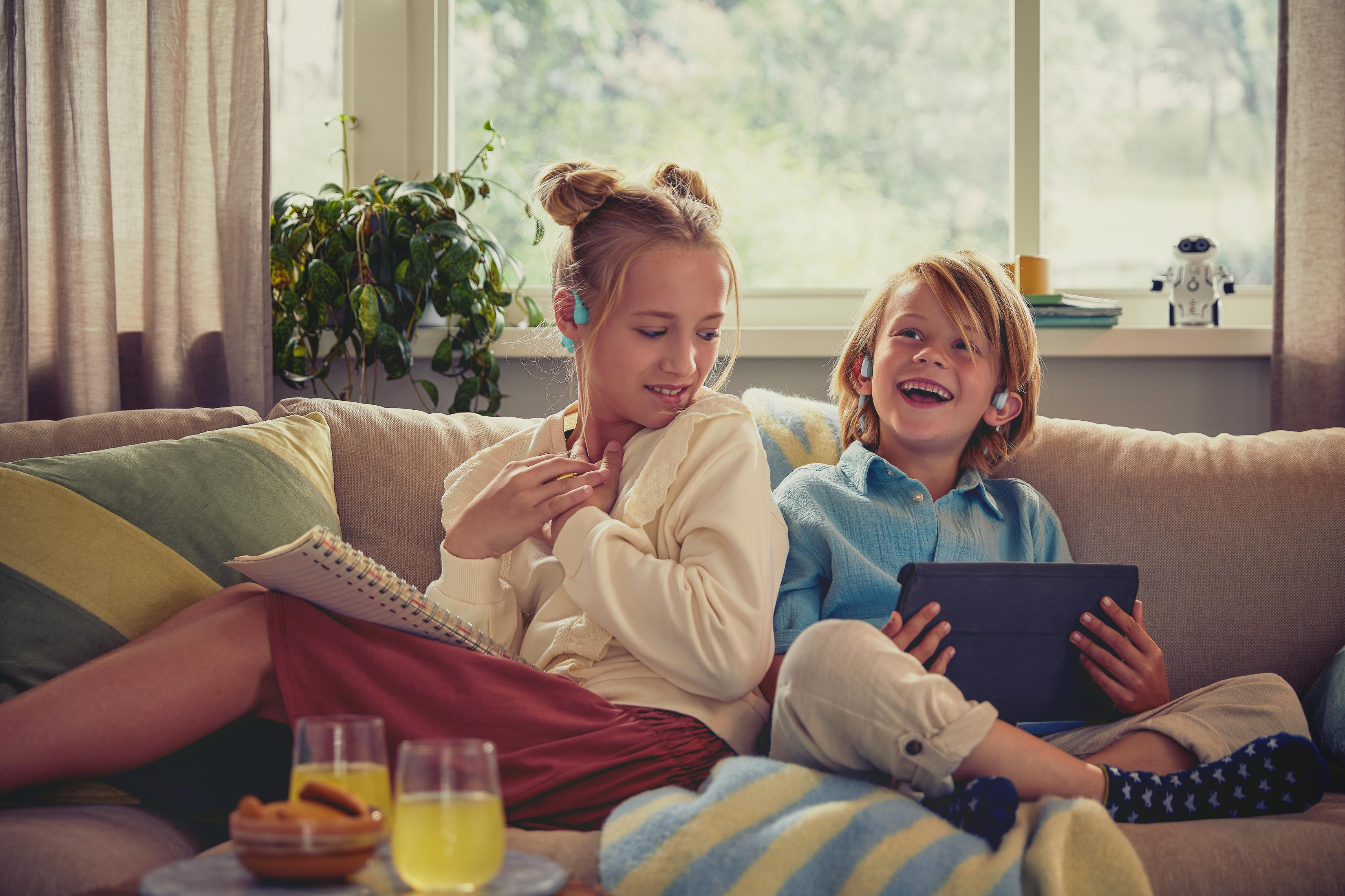 Dwoje dzieci siedzi na kanapie, uśmiecha się i ma na uszach bezprzewodowe słuchawki.