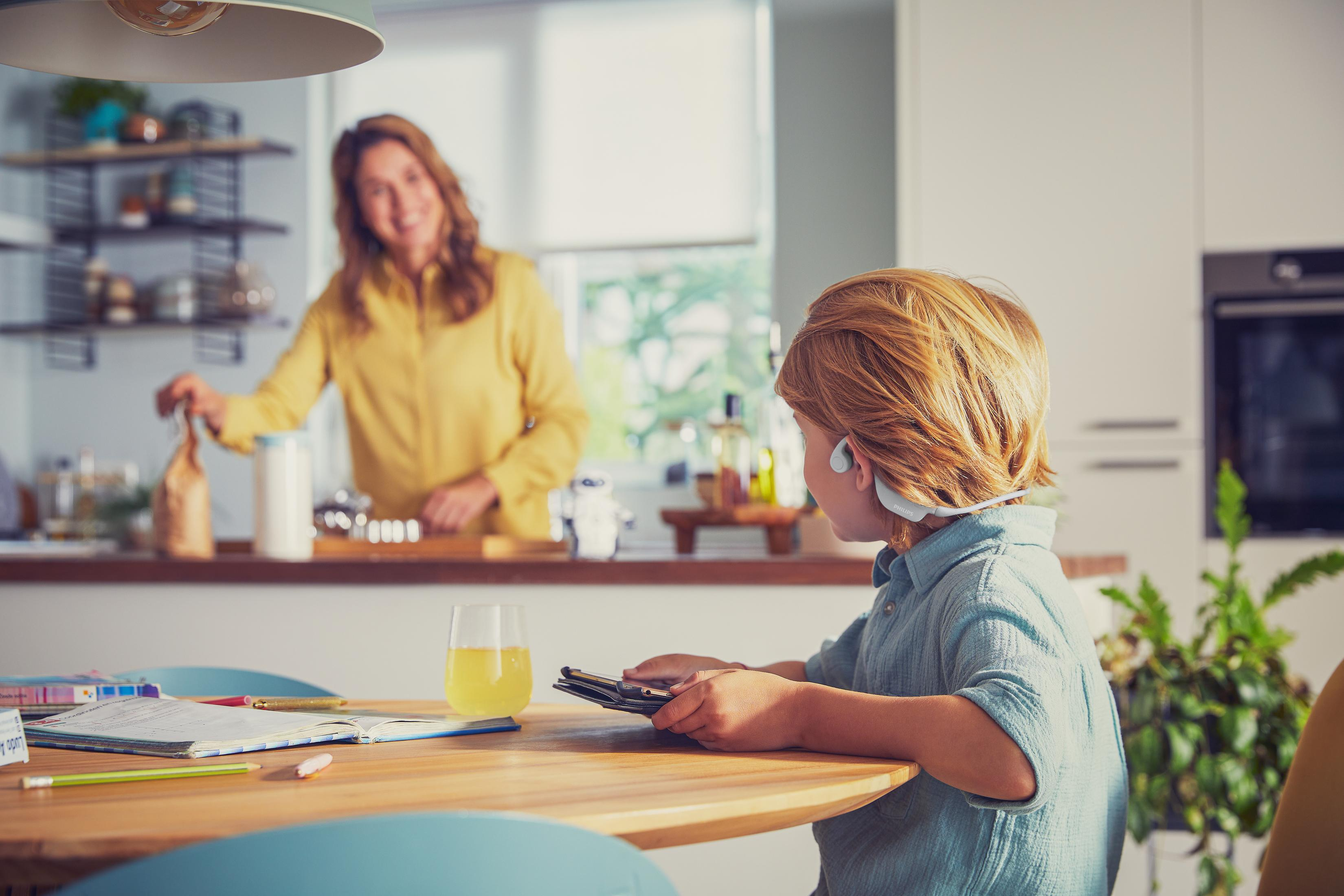 Chłopiec używa tabletu, kobieta jest w tle przy blacie kuchennym.