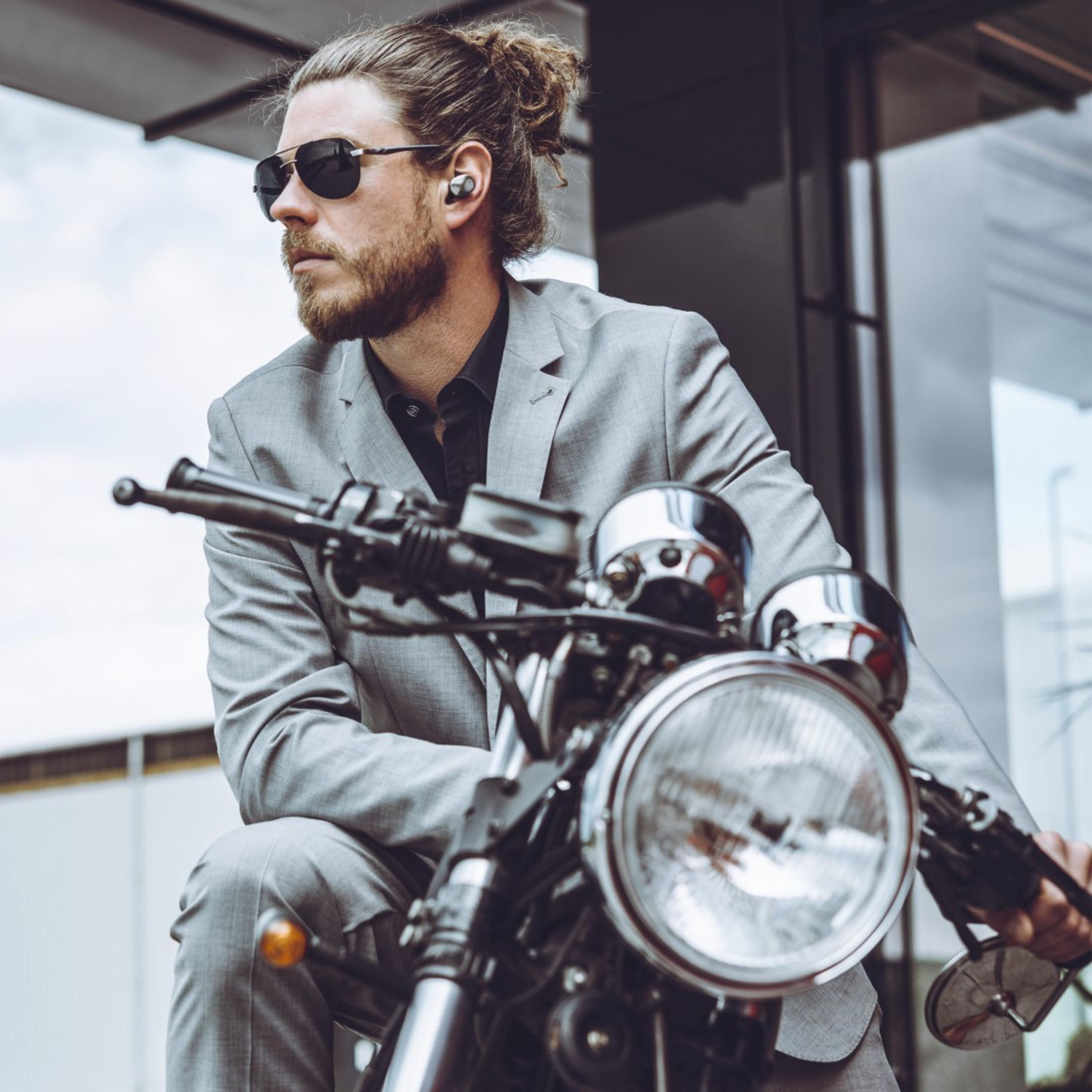 Mężczyzna w okularach przeciwsłonecznych i szarym garniturze na motocyklu, ze słuchawkami.