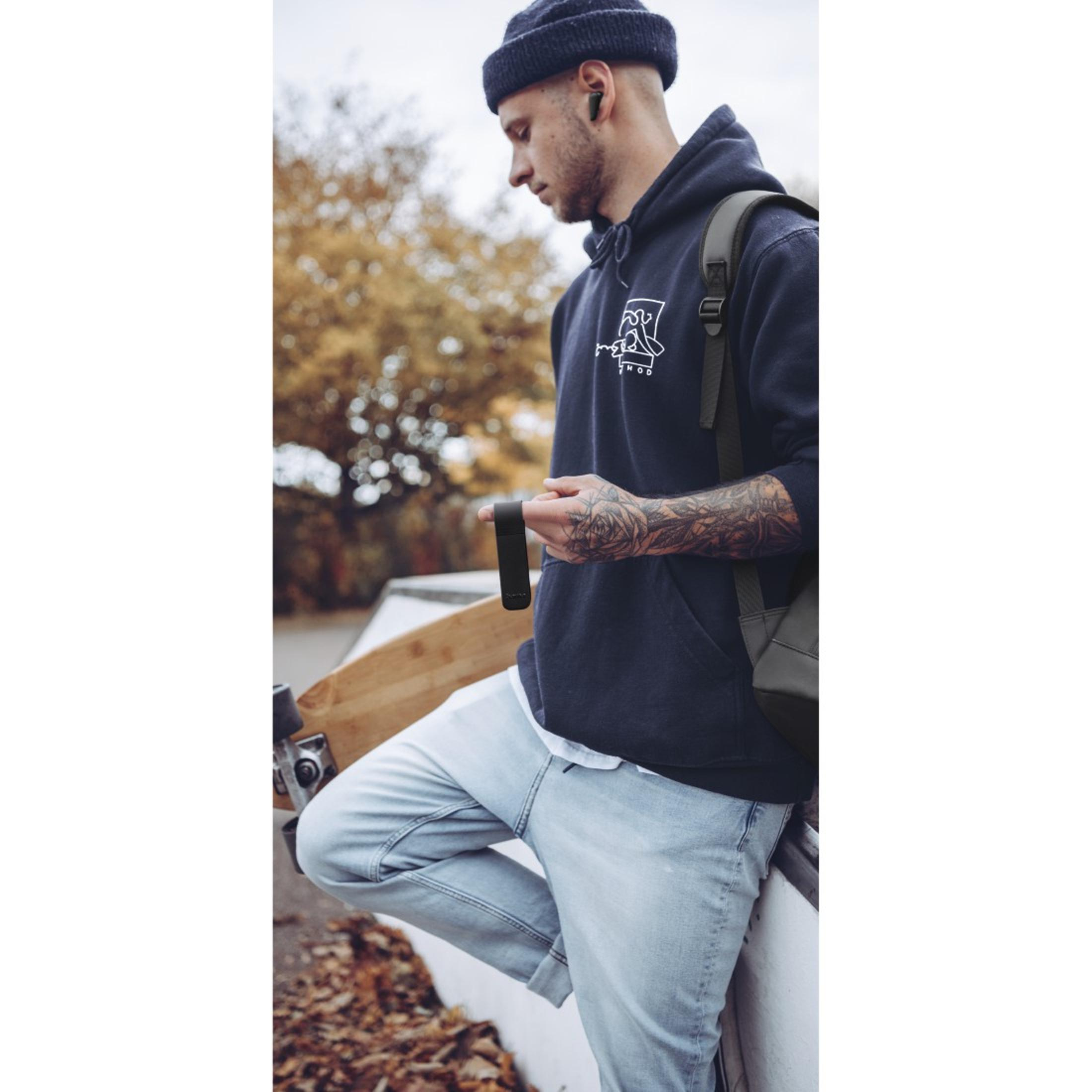 Uomo con tatuaggi, berretto, tiene una piccola custodia nera. Skatepark sullo sfondo.