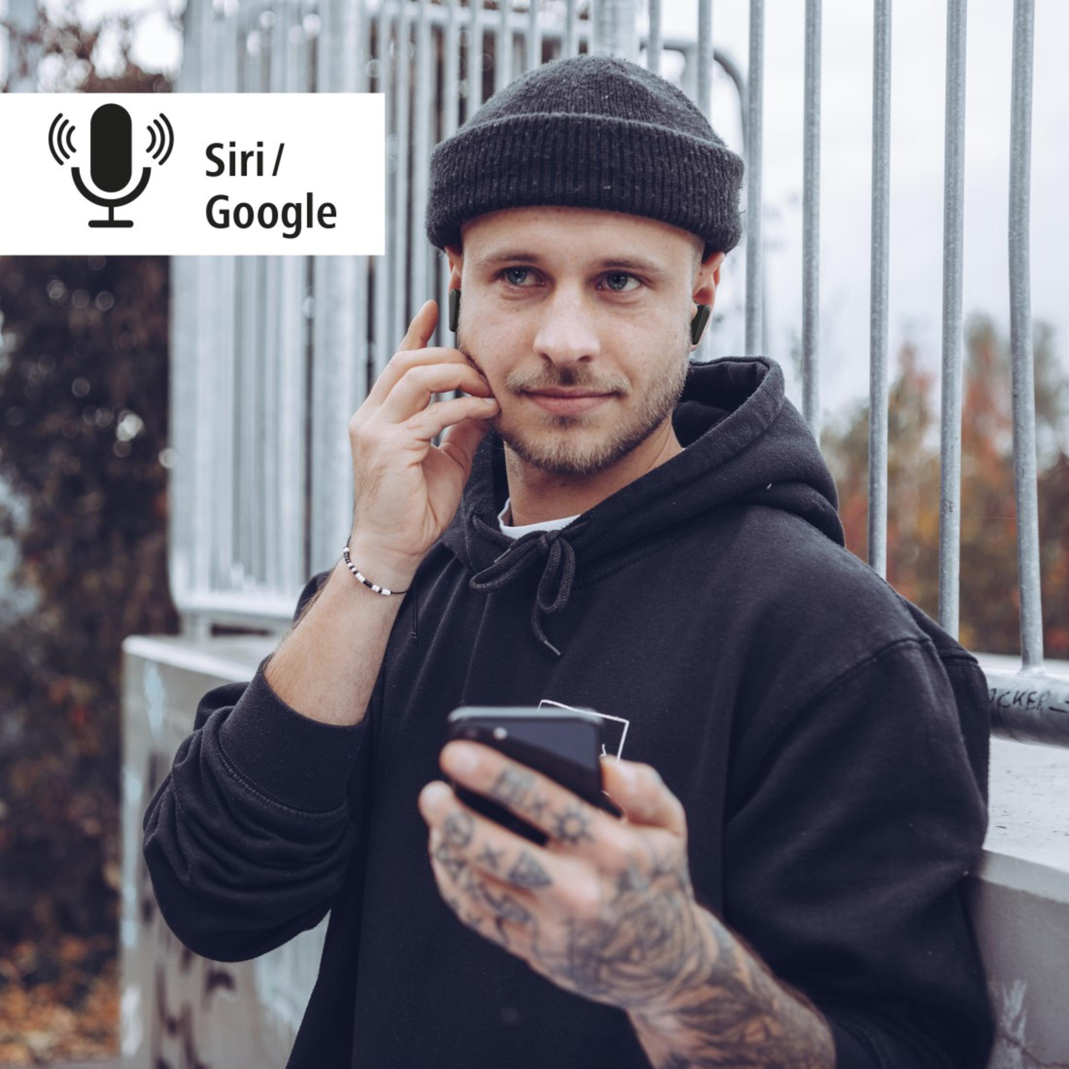 Un uomo con berretto e felpa con cappuccio neri tiene in mano un telefono, auricolari nelle orecchie. Esterno.