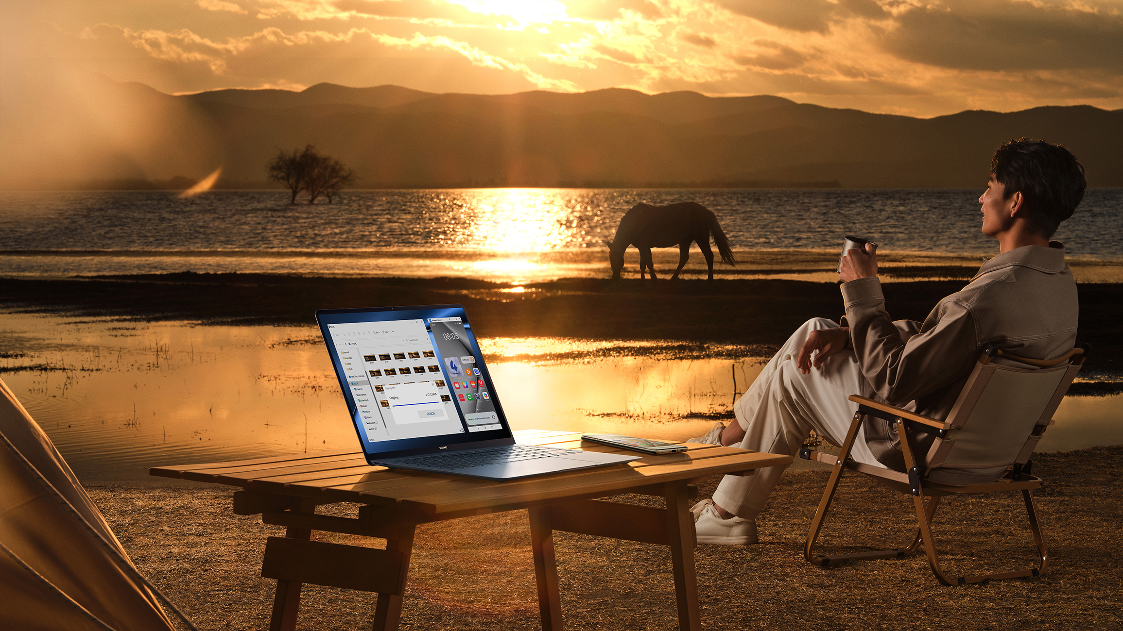 Eine Person sitzt bei Sonnenuntergang in der Nähe eines Laptops. Ein See und ein Pferd im Hintergrund. Camping.