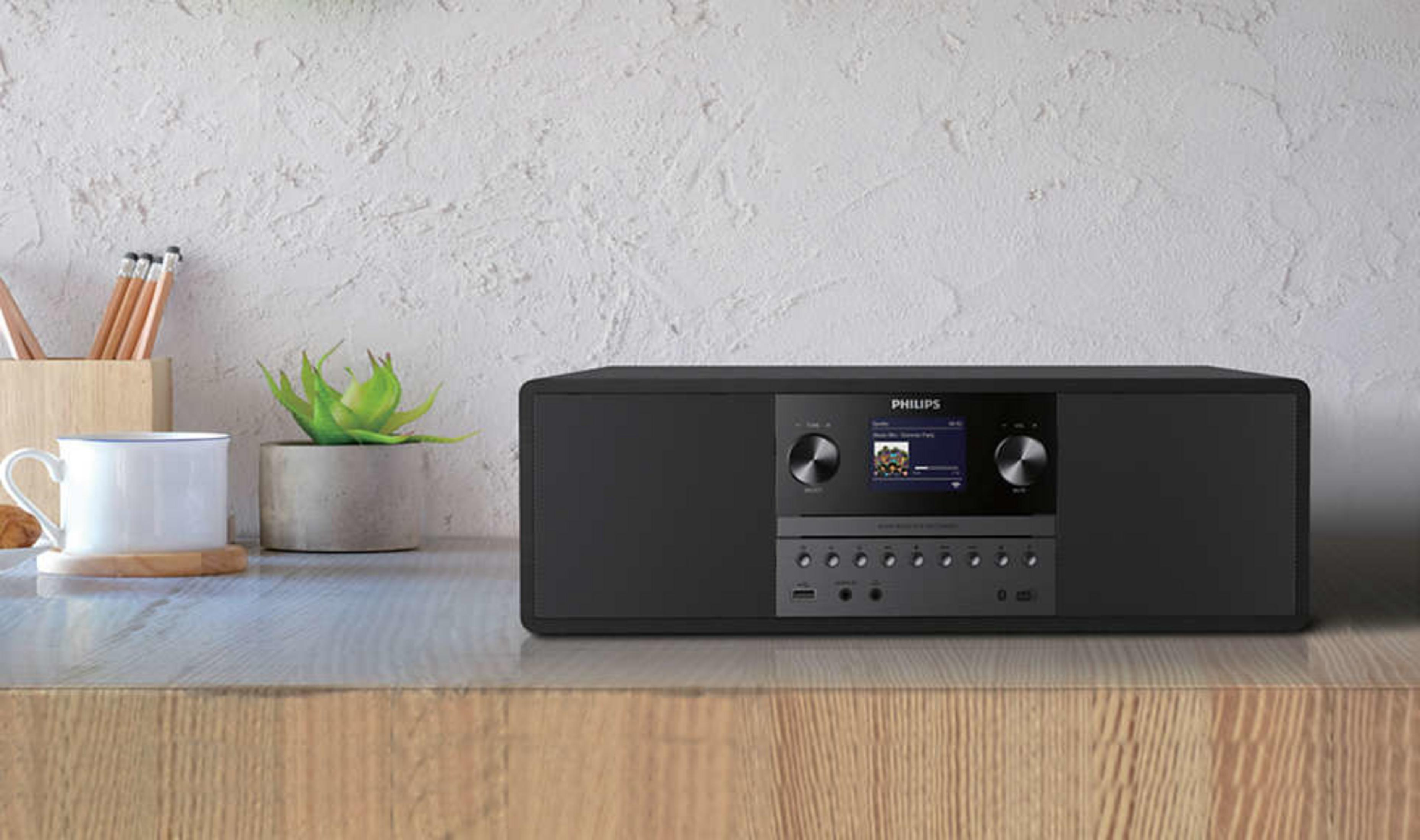 Czarny system audio Philips na drewnianej powierzchni, z rośliną, kubkiem i ołówkami w pobliżu.