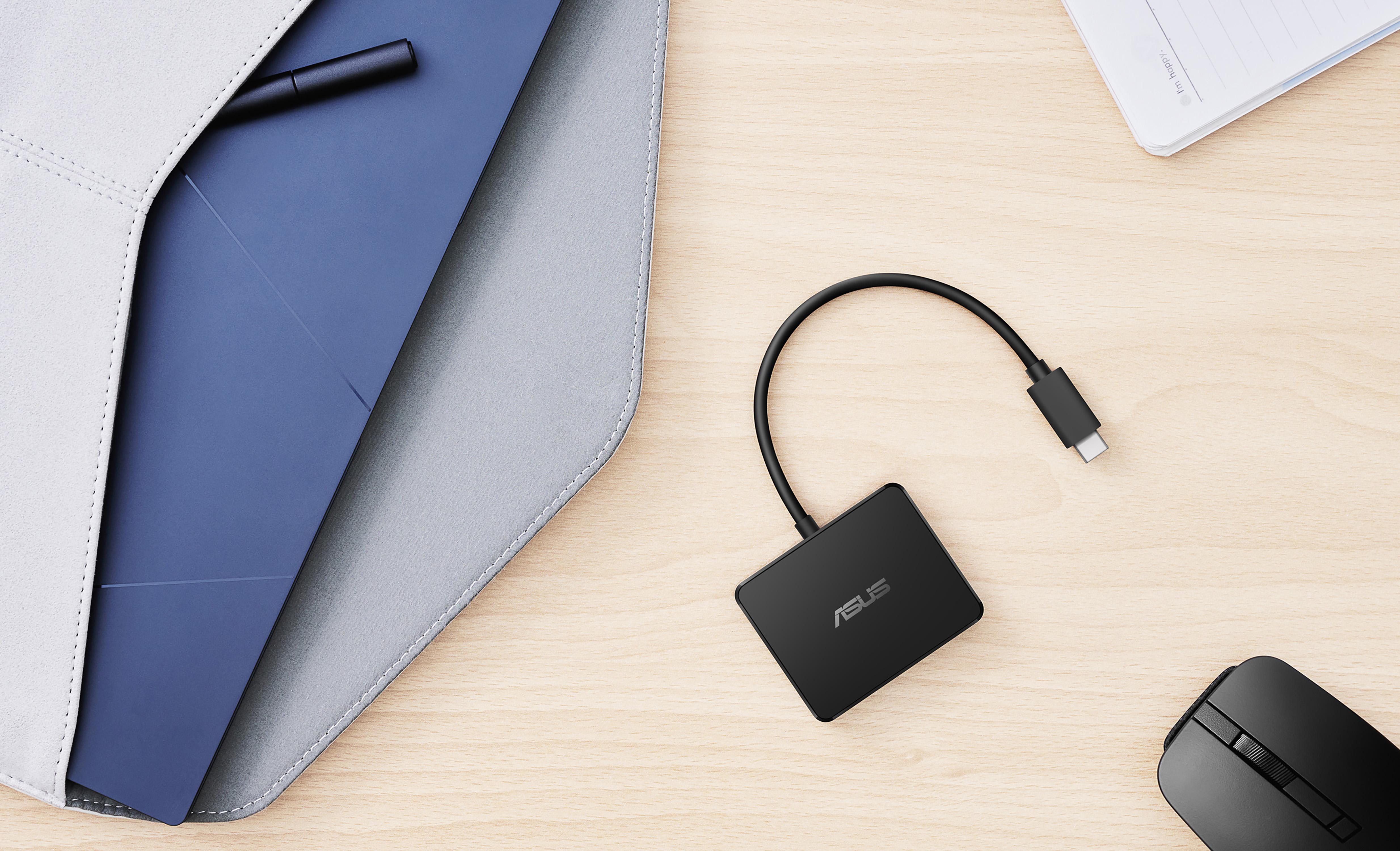 Desk setup. Laptop, pen, and mouse on wood grain. Black hub with cable. White background.