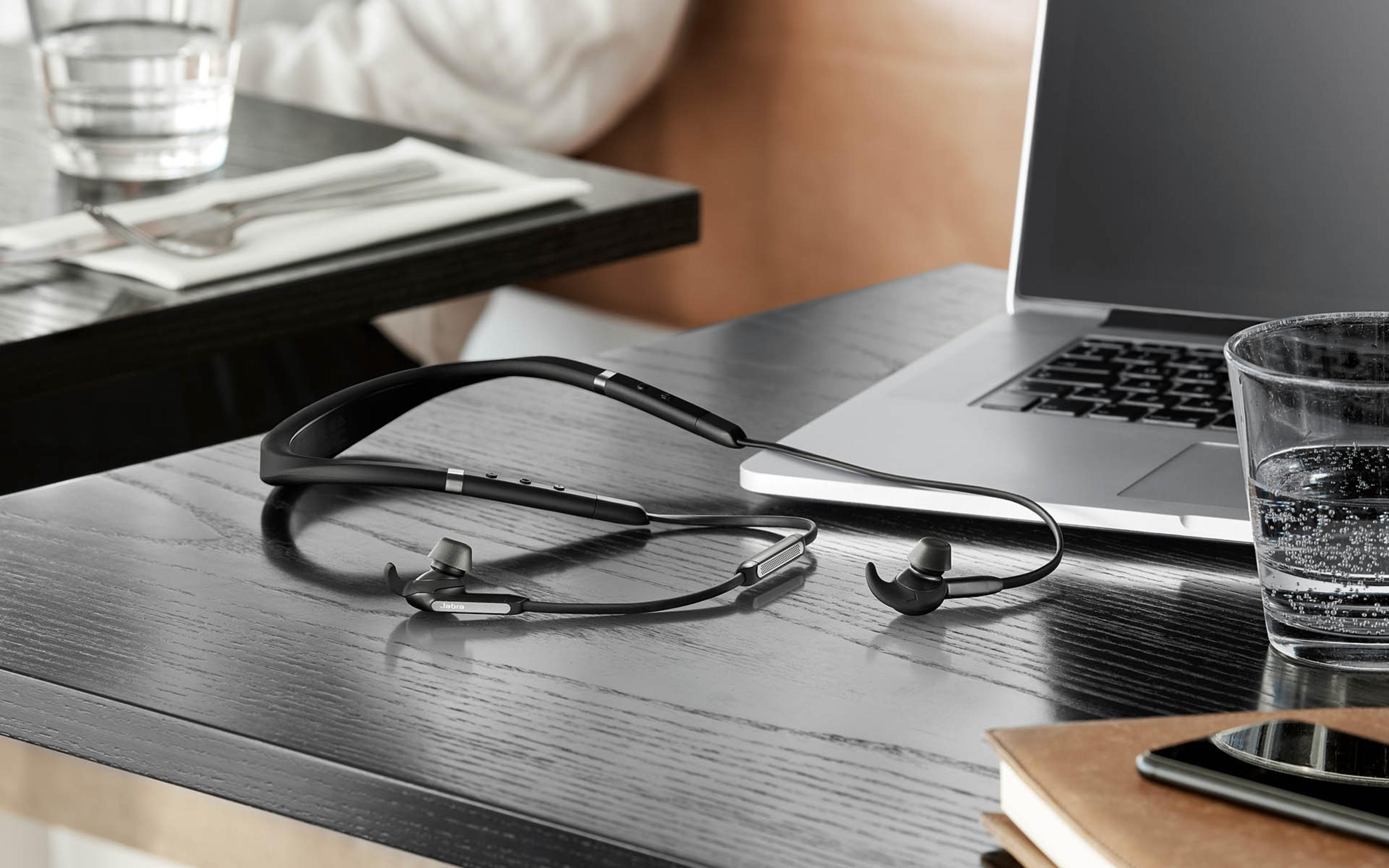 Czarne słuchawki na szyję na drewnianym stole obok laptopa i szklanki.
