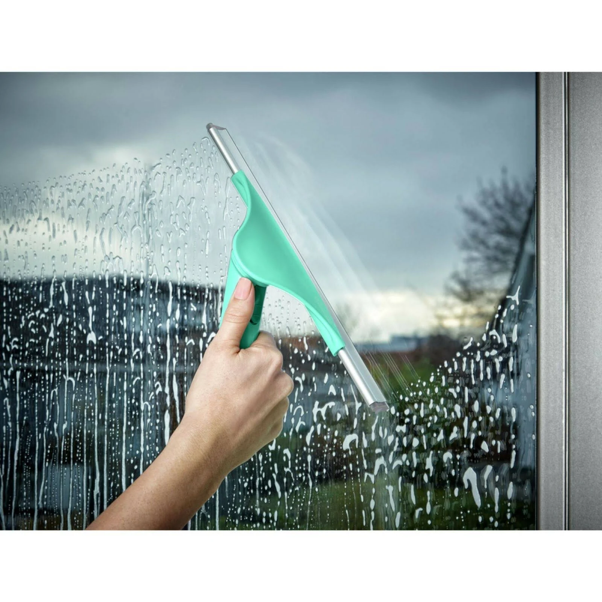 Una mano usa una espátula verde para limpiar una ventana con agua jabonosa.