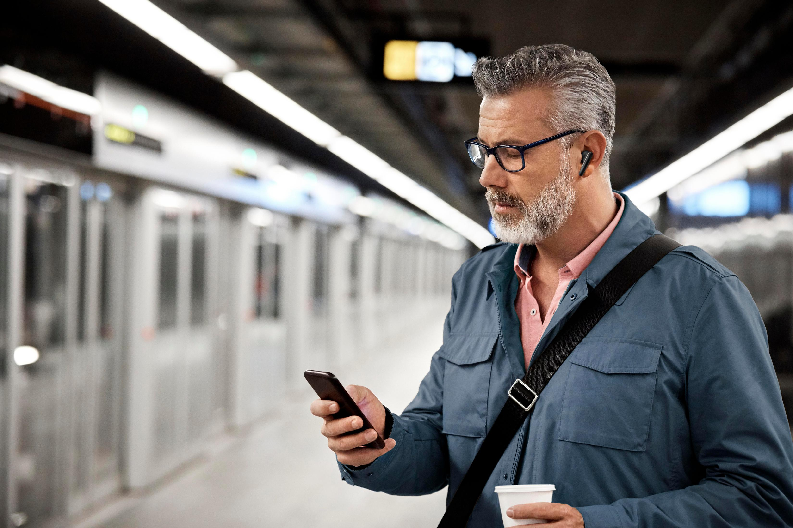 Mężczyzna w niebieskiej kurtce i okularach korzystający z telefonu na stacji metra.