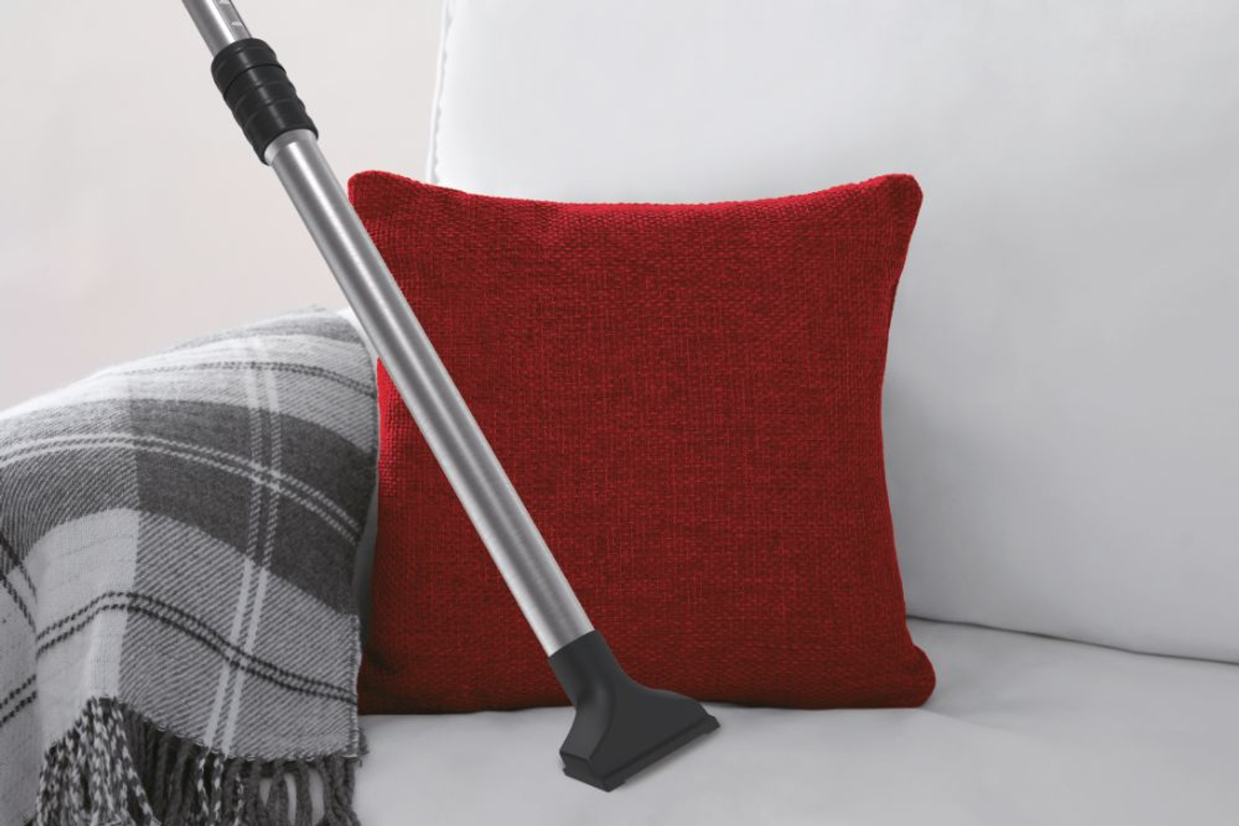 Końcówka odkurzacza w pobliżu czerwonej poduszki i kratkowanego koca na białej sofie.