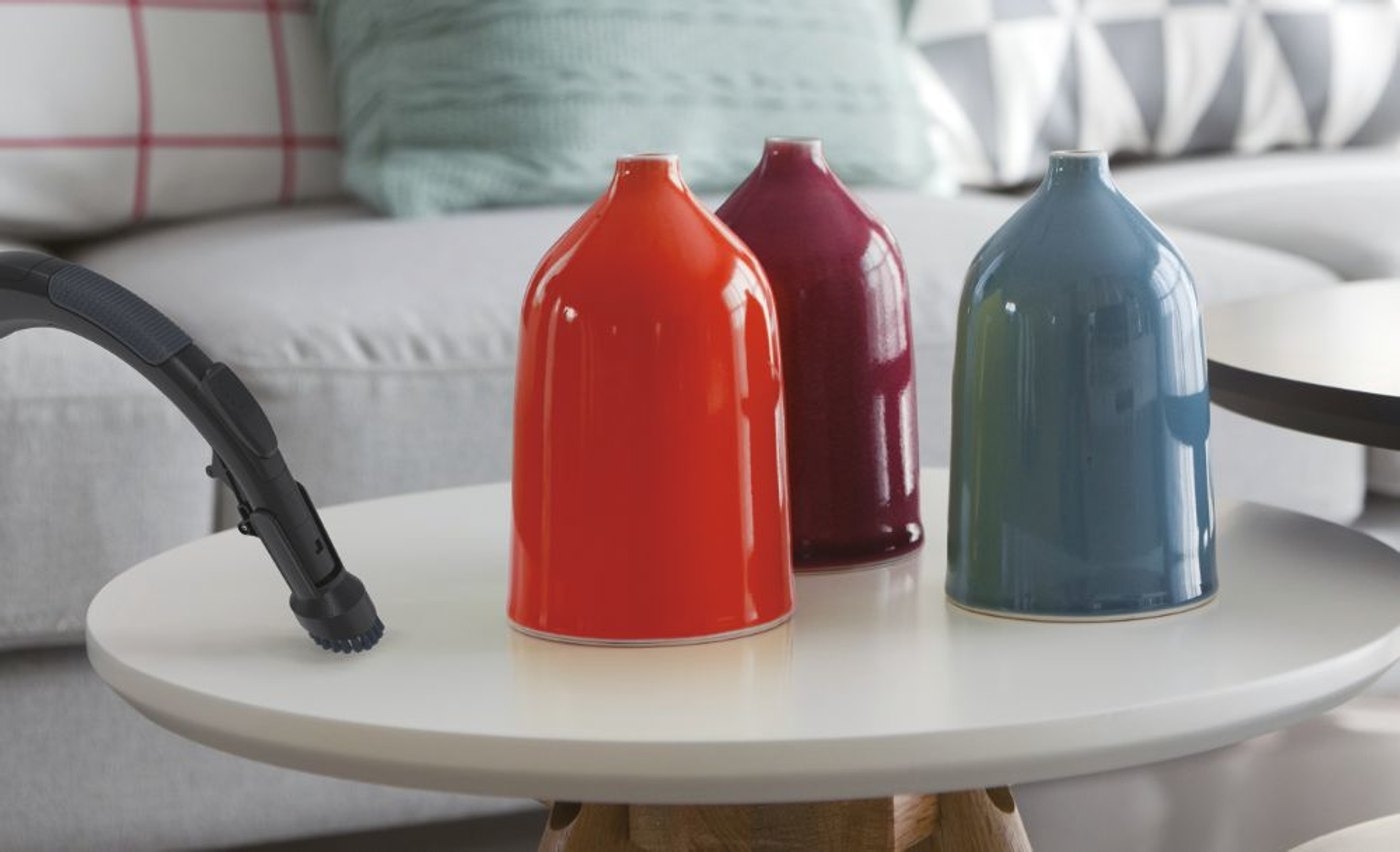 Trzy kolorowe wazony, czerwony, fioletowy i niebieski, na białym stole. Z lewej strony odkurzacz.
