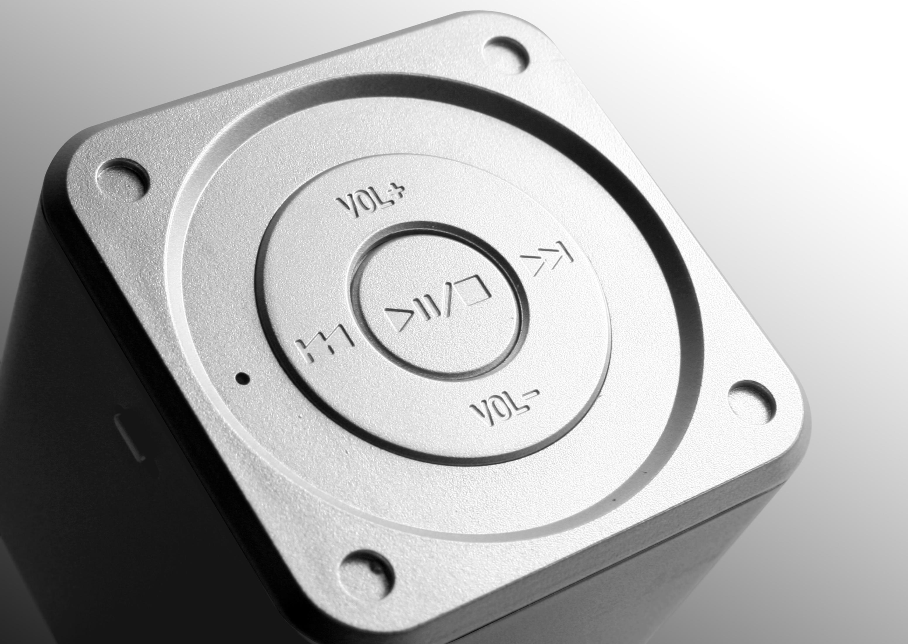 Zbliżenie srebrnego głośnika z przyciskami sterowania. Przyciski głośności i odtwarzania.