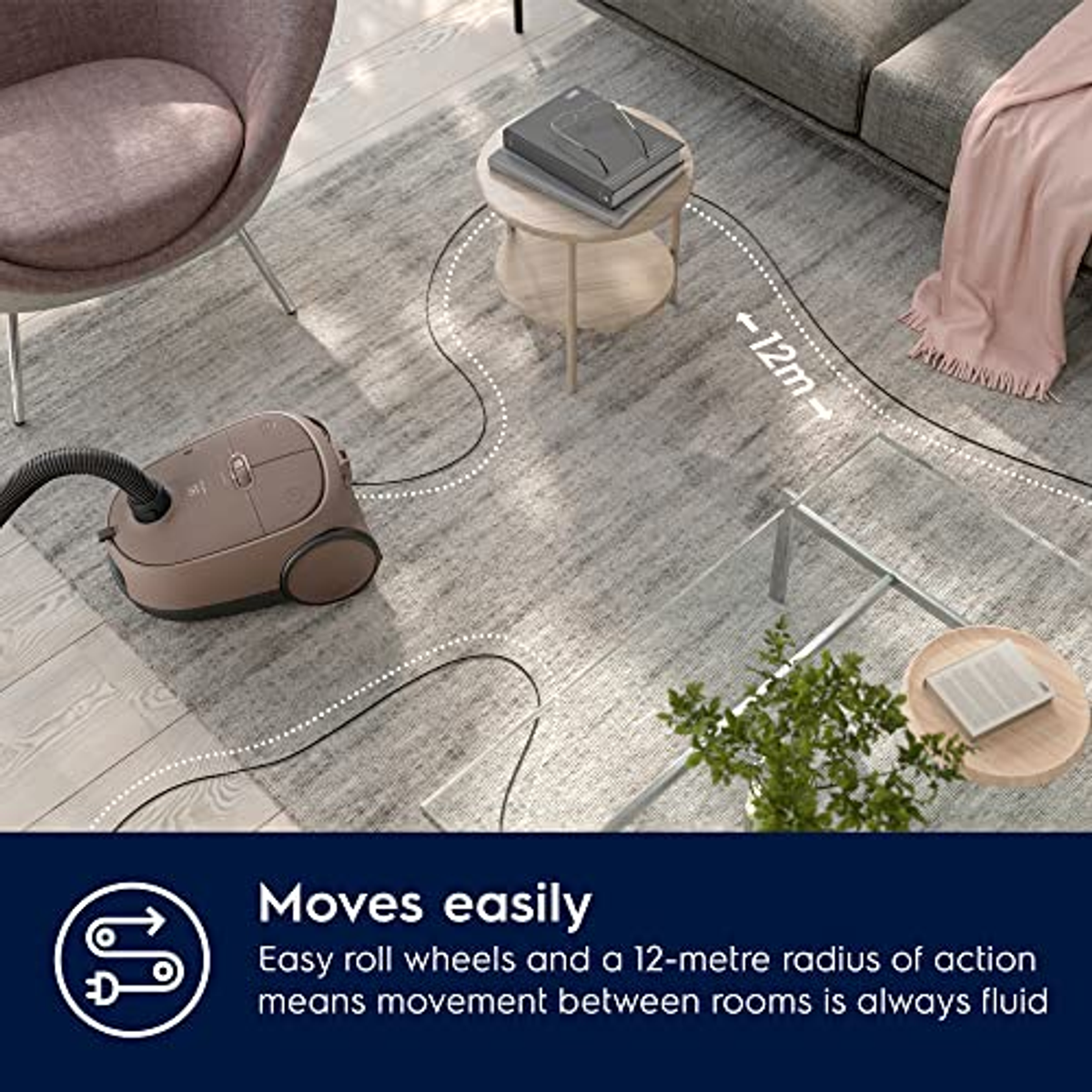 Odkurzacz na dywanie, wąż po lewej, stół z książkami i krzesło w tle.