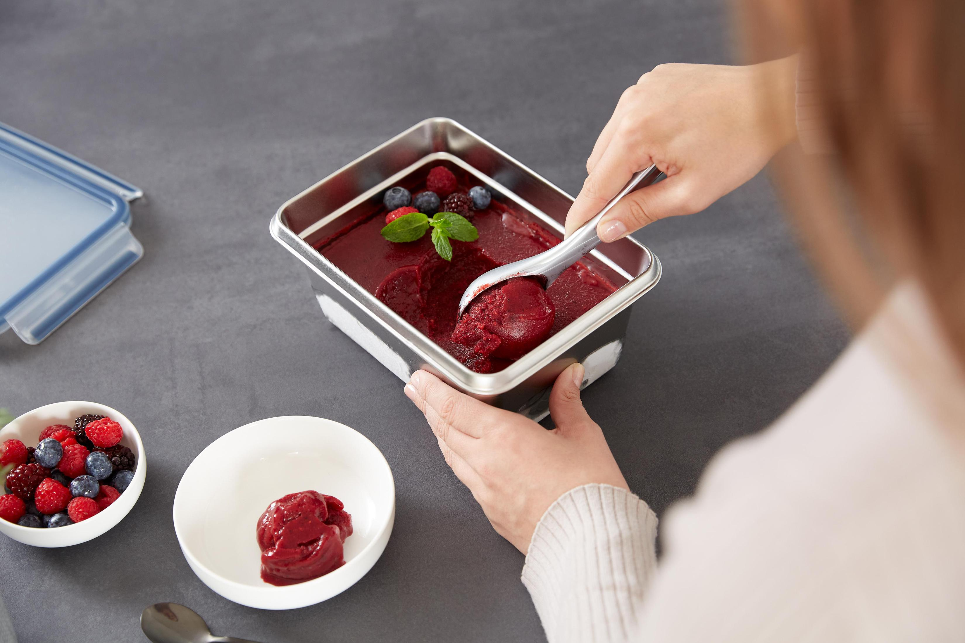 Eine Person schöpft rotes Sorbet aus einem Behälter. Beeren sind im Hintergrund.