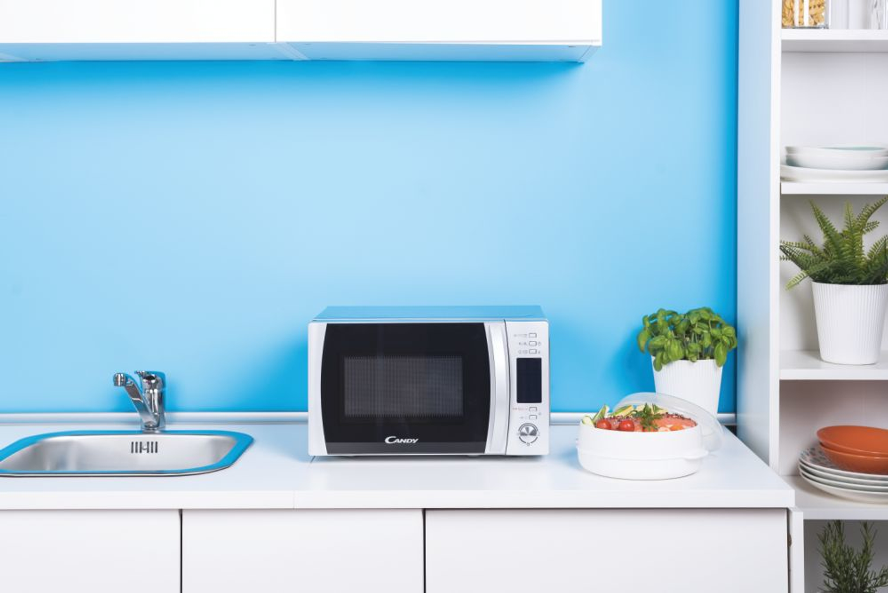 Una cocina blanca con microondas, fregadero, plantas y un recipiente con comida en la encimera.
