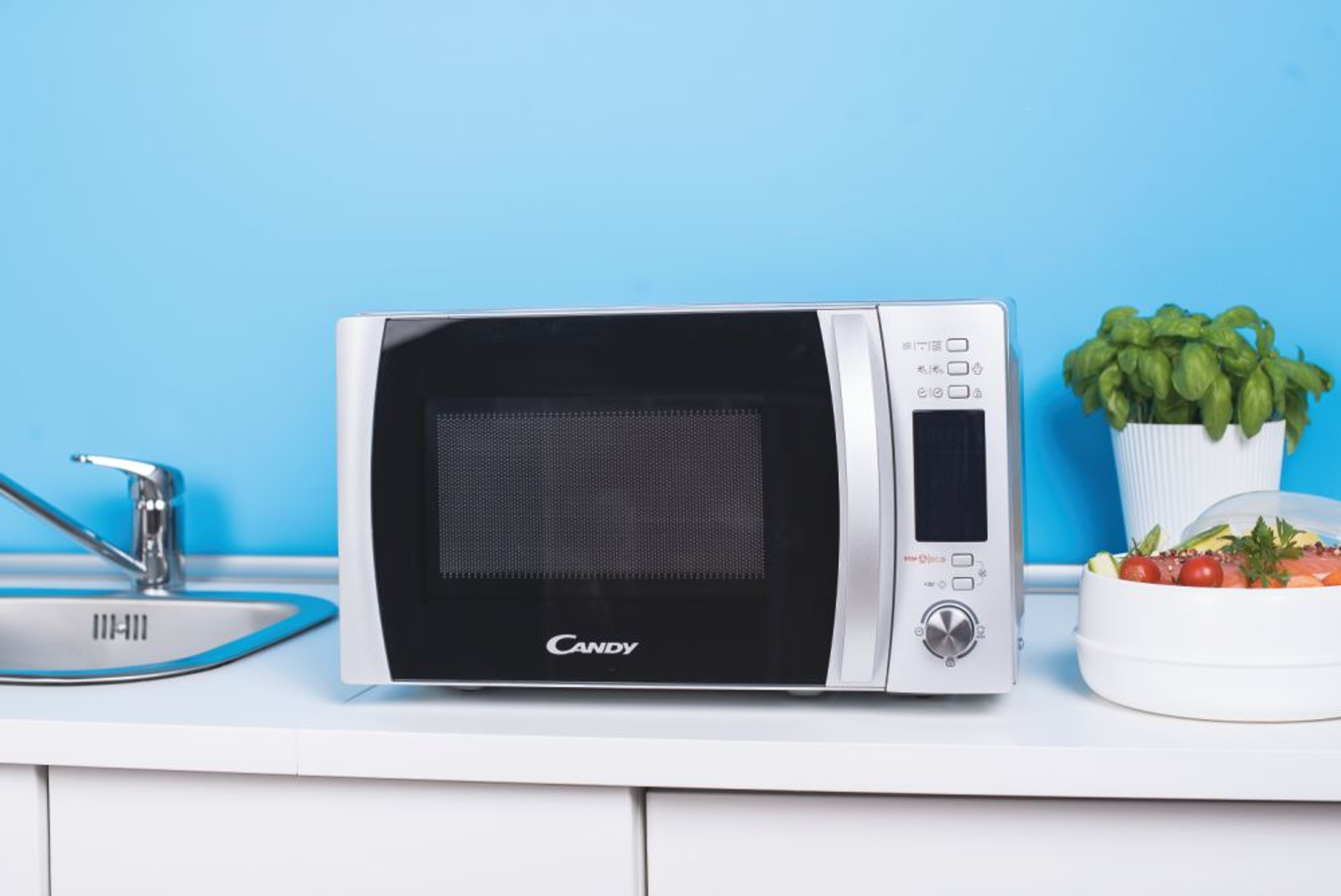 Un horno microondas Candy plateado sobre una encimera blanca con un fregadero y comida.