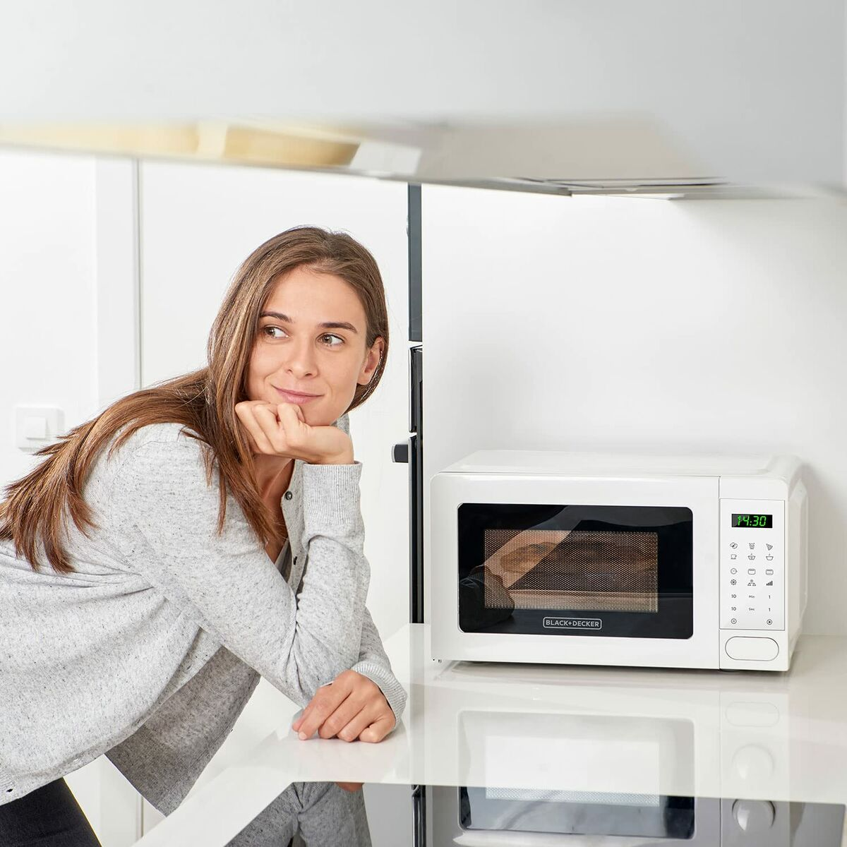Kobieta opiera się o biały blat, obok biało-czarnej kuchenki mikrofalowej Black+Decker.