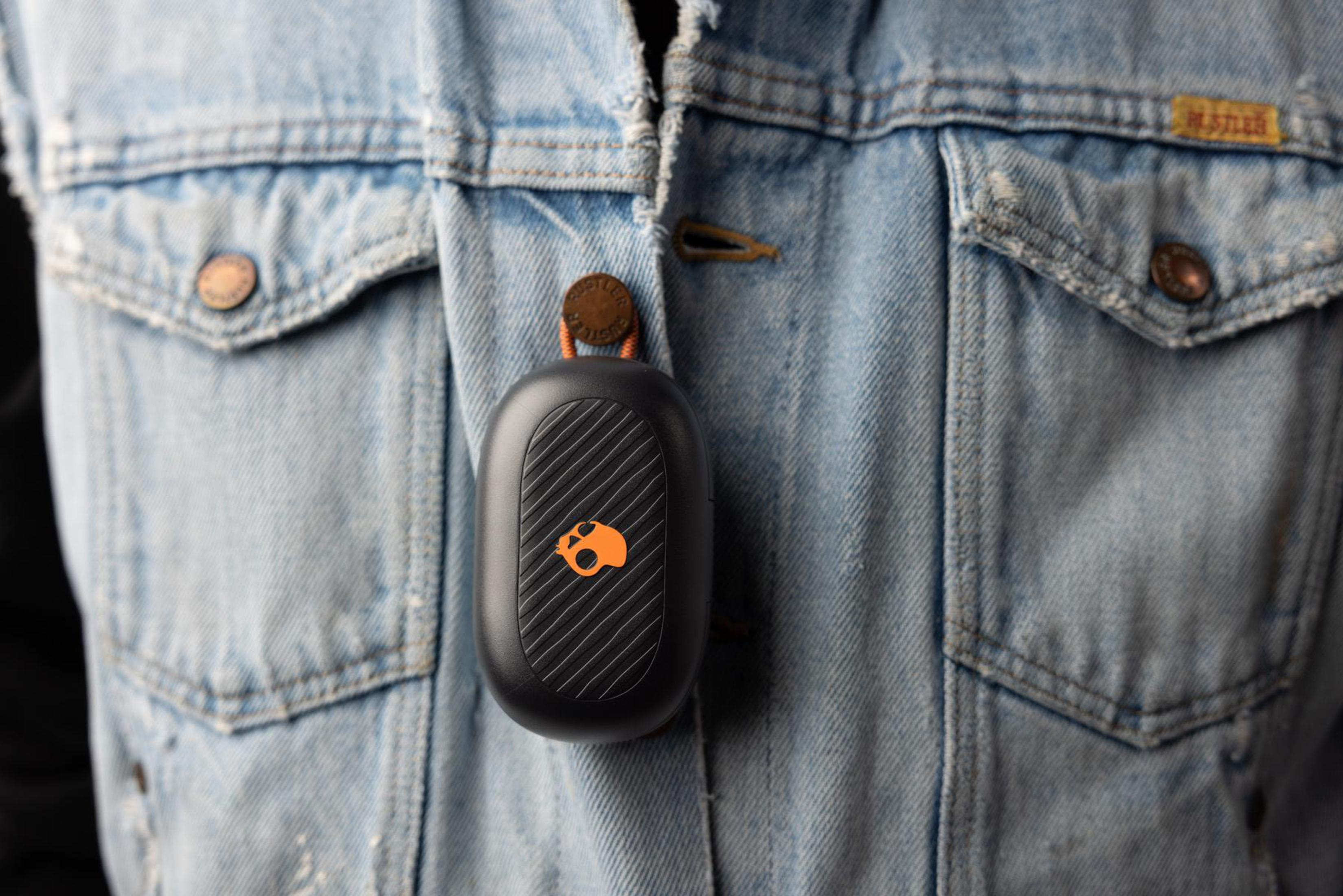 Czarny i szary futerał na słuchawki przymocowany do dżinsowej kurtki. Futerał ma logo i pomarańczowe detale.