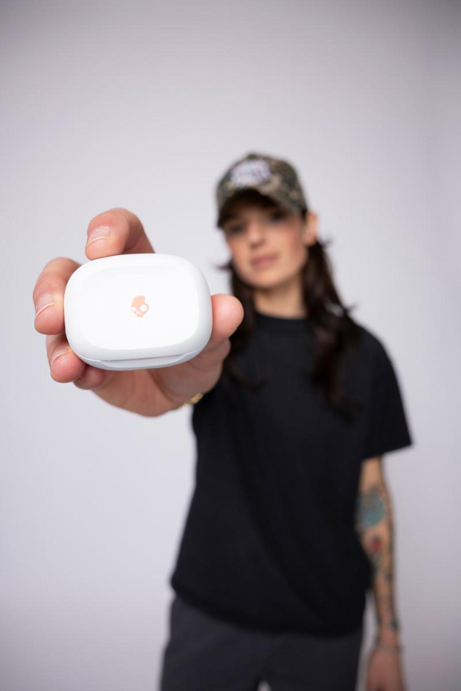 Osoba trzymająca białe etui na słuchawki. Osoba ma na sobie czarną koszulę i czapkę moro. Białe tło.