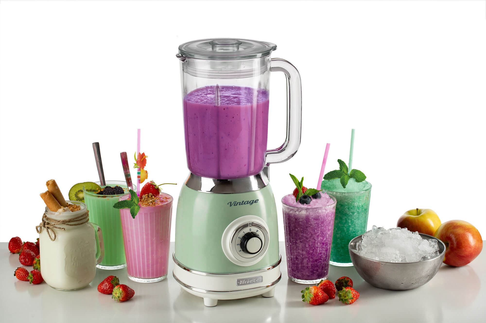 Blender met een groene basis en een glazen pot. Verschillende smoothies en ingrediënten op tafel.