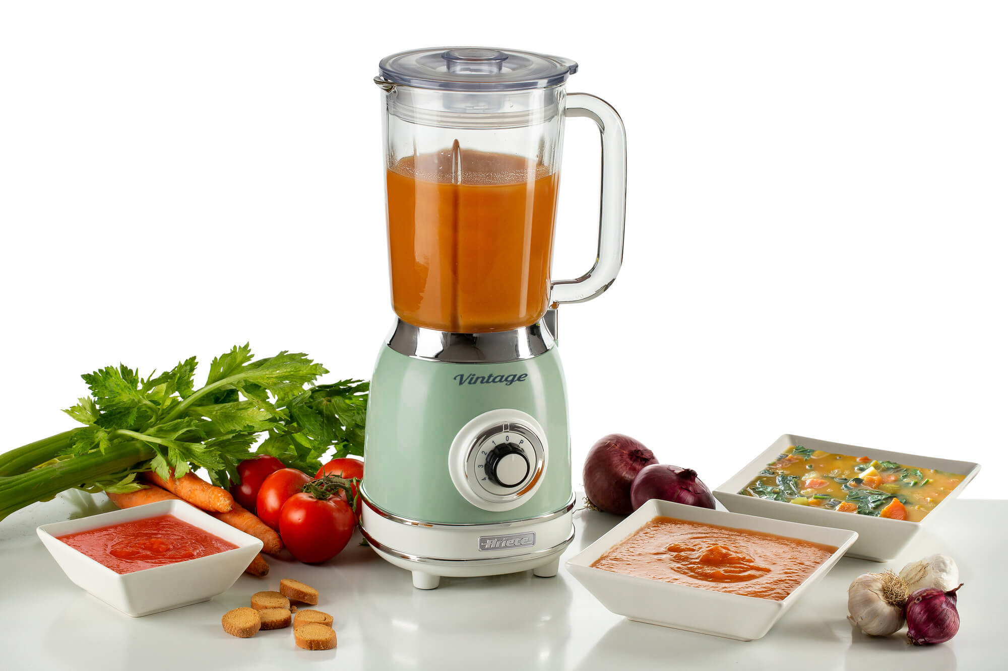 Een blender met oranje vloeistof, groenten en kommen met eten.