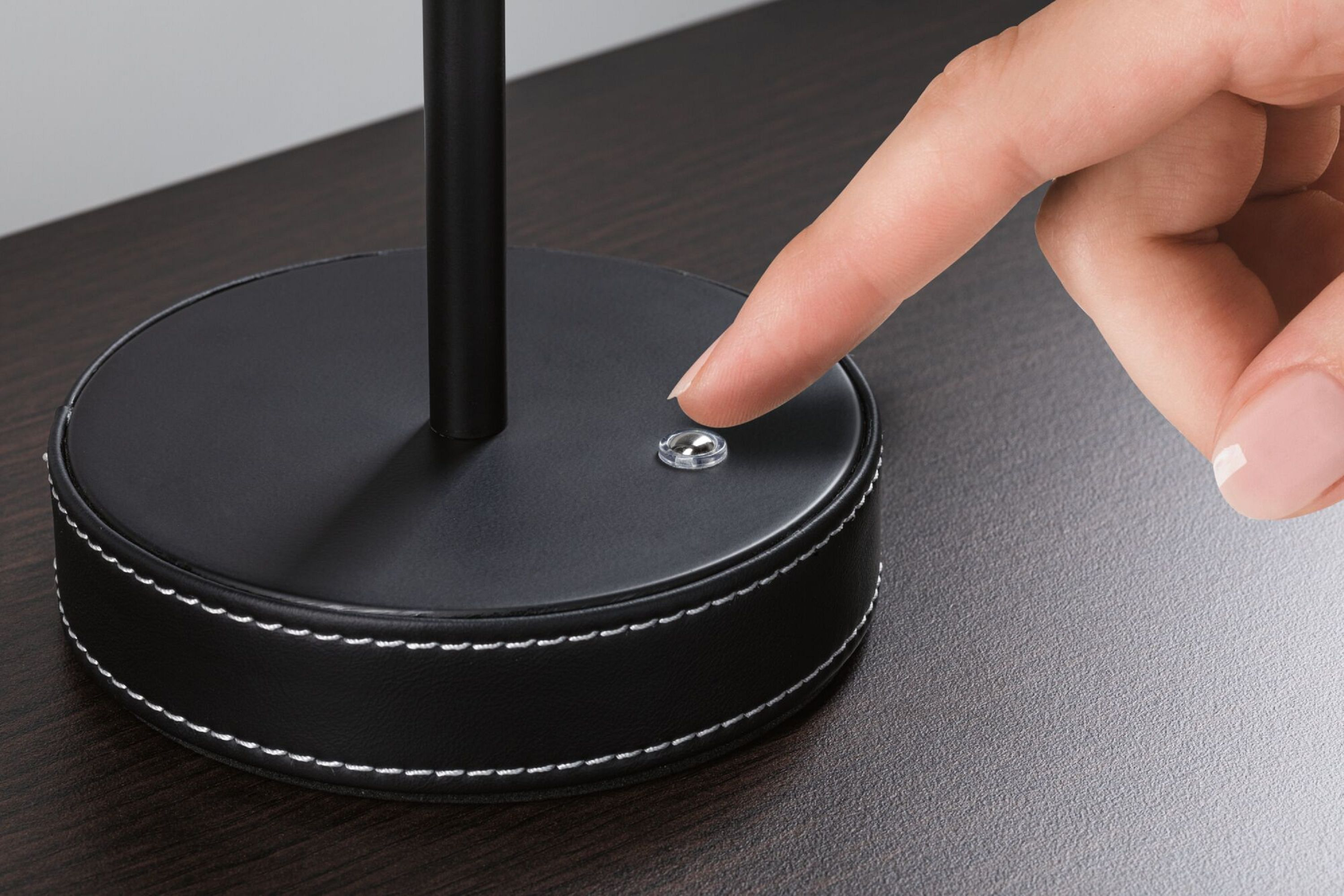 Dłoń naciska włącznik na czarnej podstawie lampy. Ciemna powierzchnia drewna.