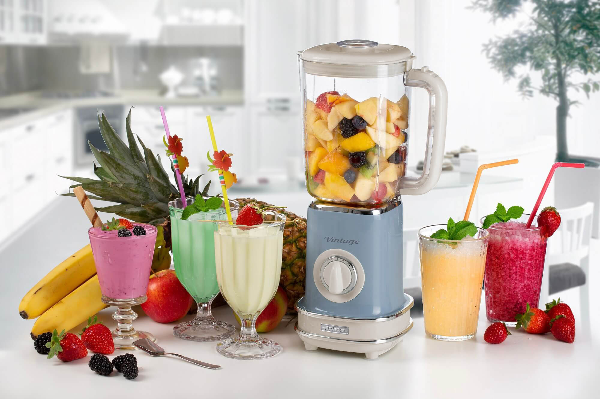 Frullatore con frutta all'interno. Diversi bicchieri di frullati e frutta sul tavolo.