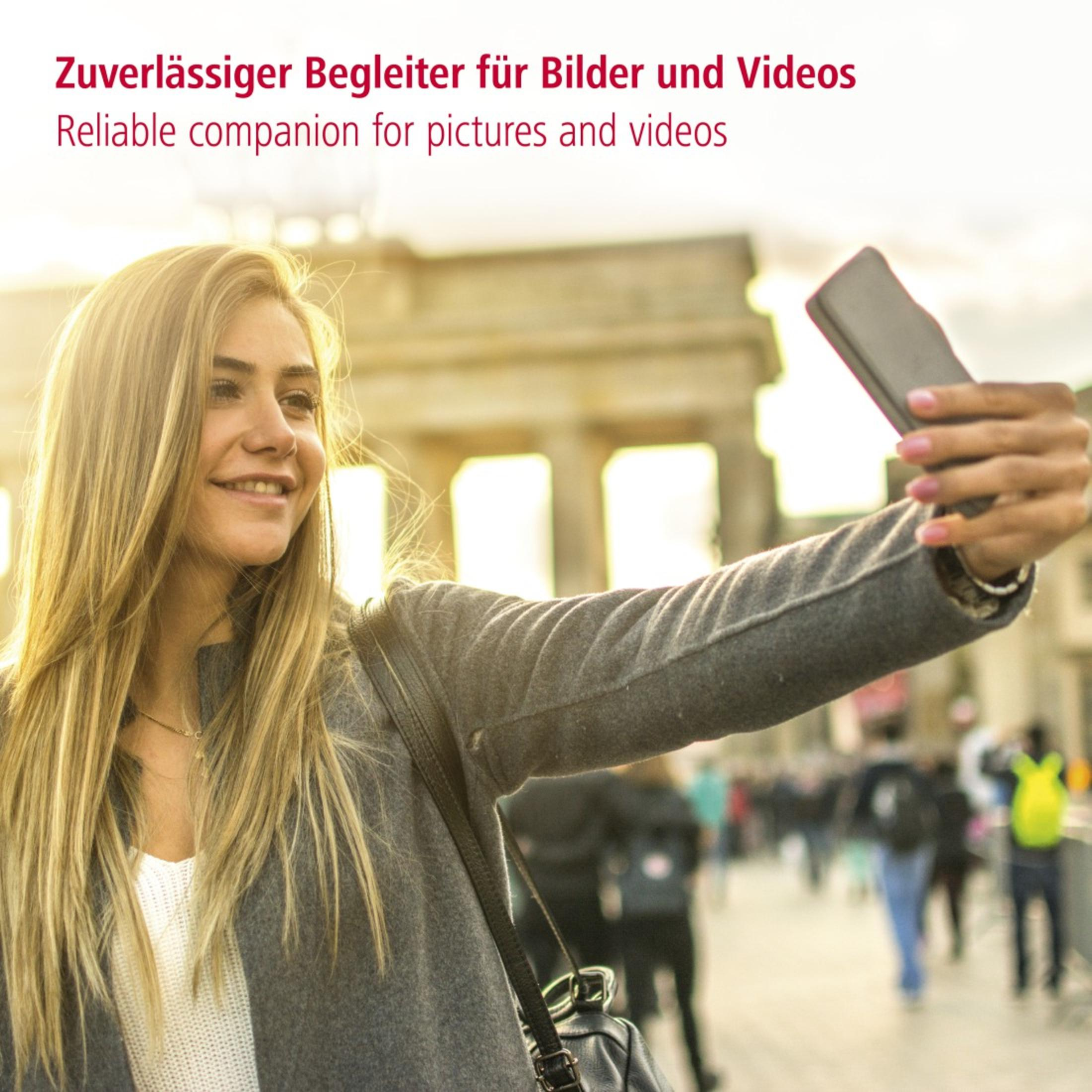 Frau macht ein Selfie mit einem Smartphone vor einem verschwommenen Hintergrund eines Gebäudes und Personen.