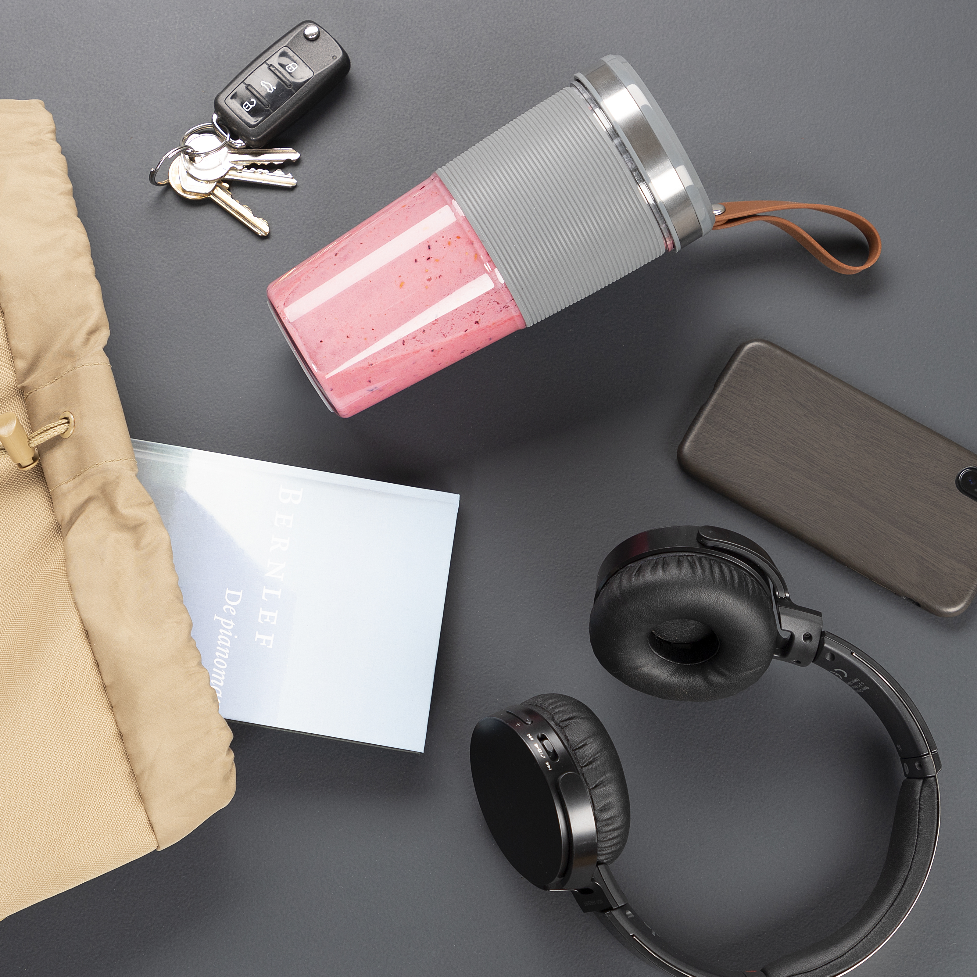 Szara powierzchnia z kubkiem smoothie, kluczami, telefonem, słuchawkami i torbą. Przedmioty ułożone swobodnie.