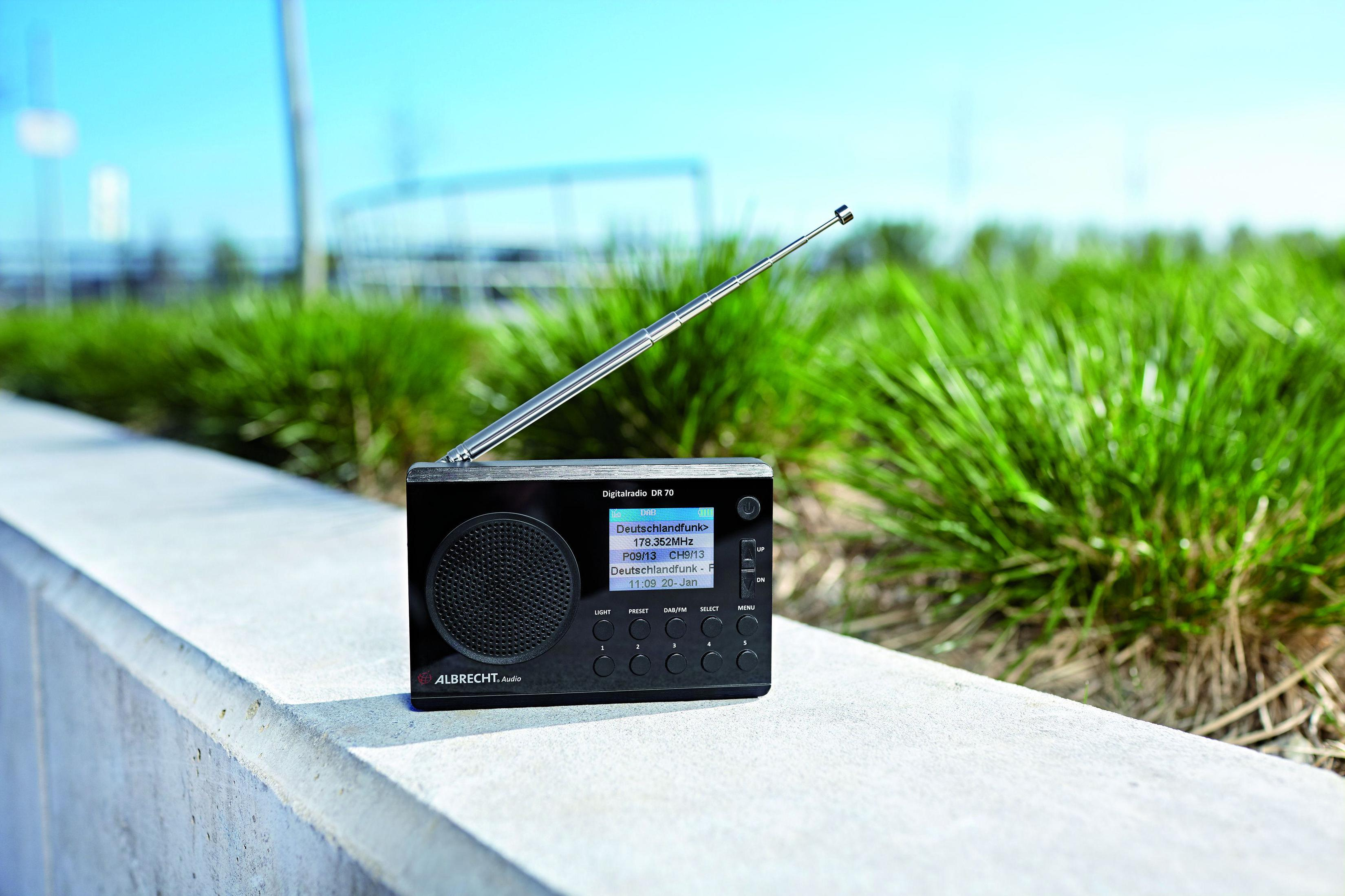 Radio nera su un davanzale, antenna in alto, testo sullo schermo, con cespugli verdi sullo sfondo.
