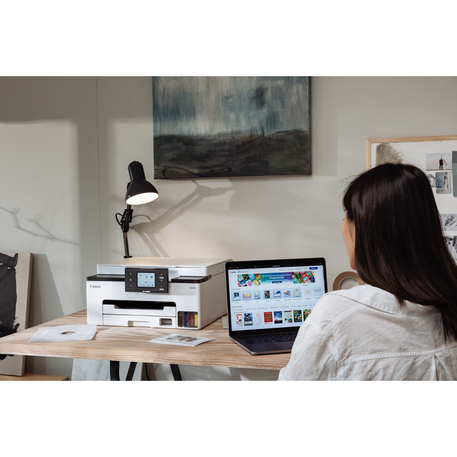 Kobieta używa laptopa obok drukarki, z obrazem na ścianie.