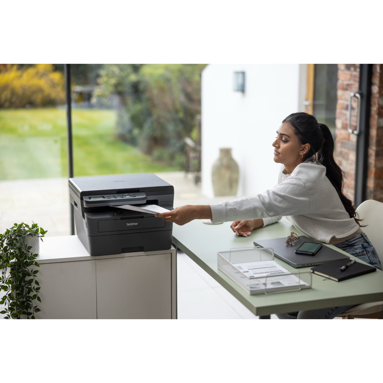 Kobieta wkłada kartkę do drukarki Brother, z laptopem i innymi artykułami biurowymi na biurku.