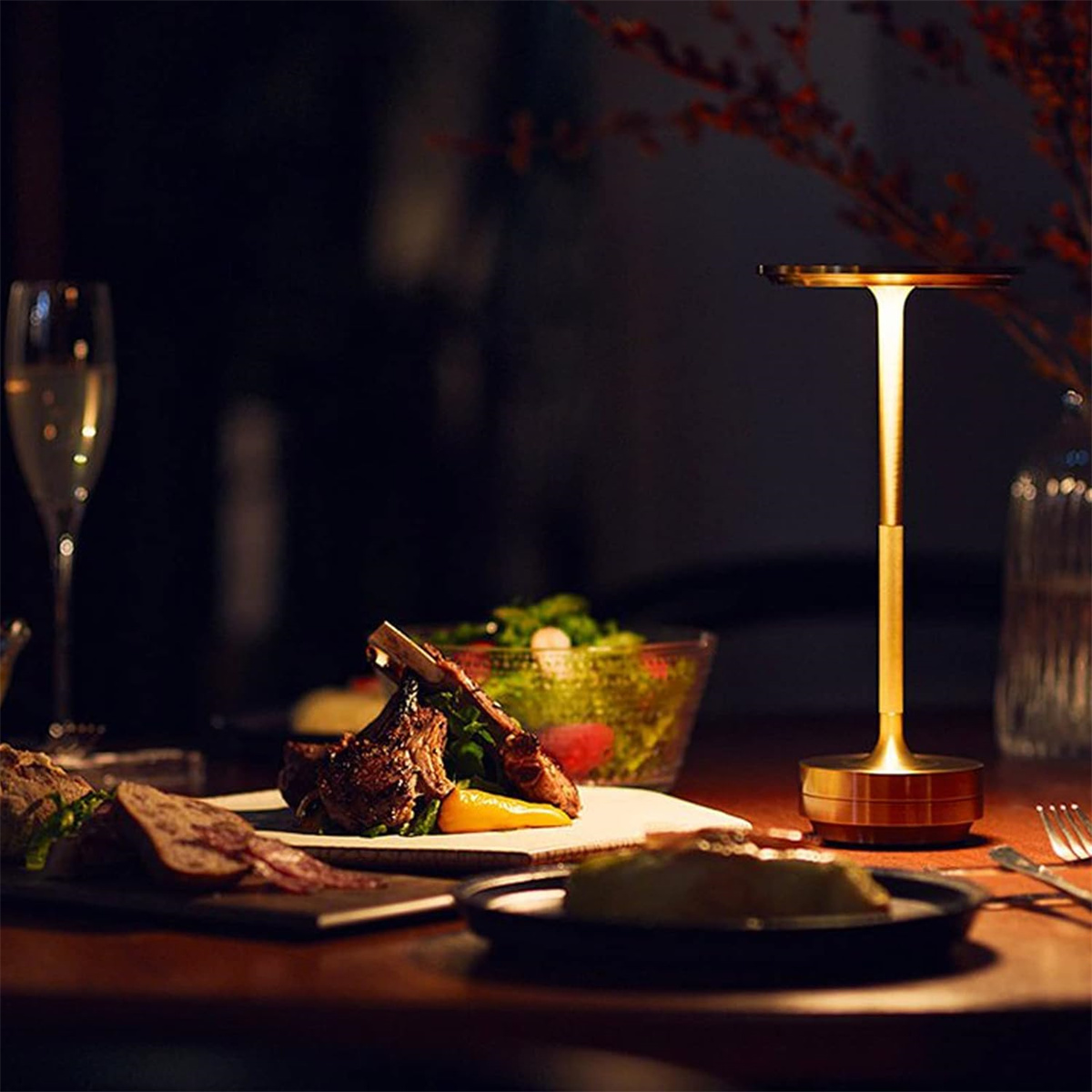 Tisch mit Lammkoteletts, goldener Lampe, Weinglas und Salat. Dunkler, verschwommener Hintergrund.