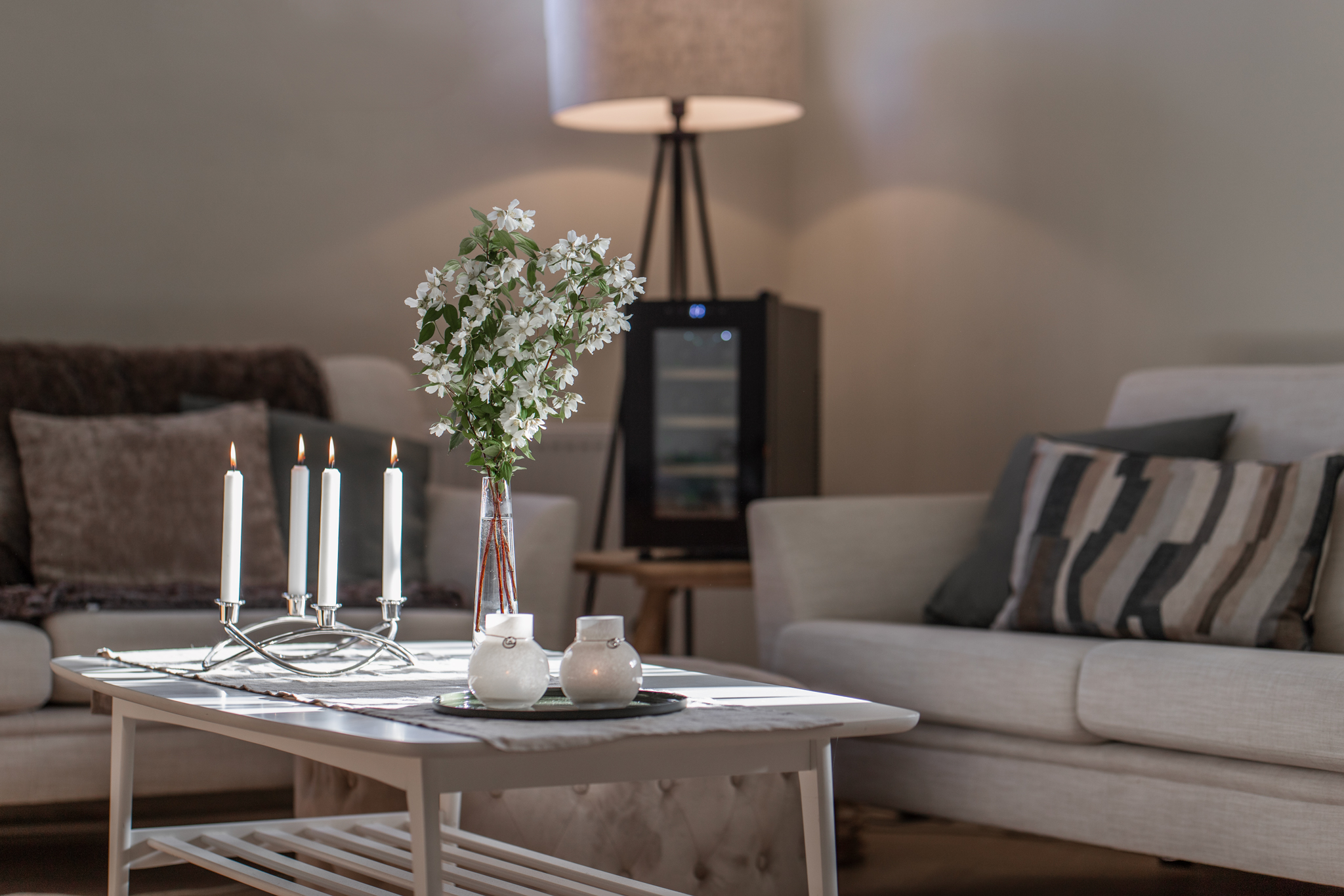 Wohnzimmerszene mit weißem Tisch mit Kerzen und Blumen. Sofas und eine Lampe sind im Hintergrund.