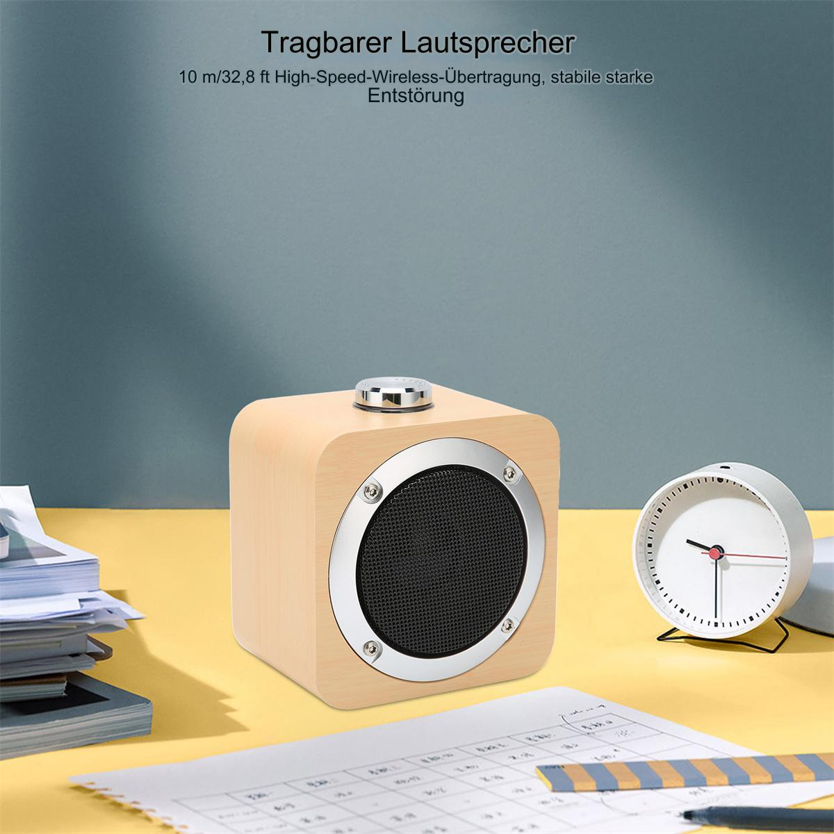 Ein kleiner, hölzerner Würfel-Lautsprecher mit einem silbernen Ring um den Lautsprecher. Eine Uhr und Papiere sind in der Nähe.