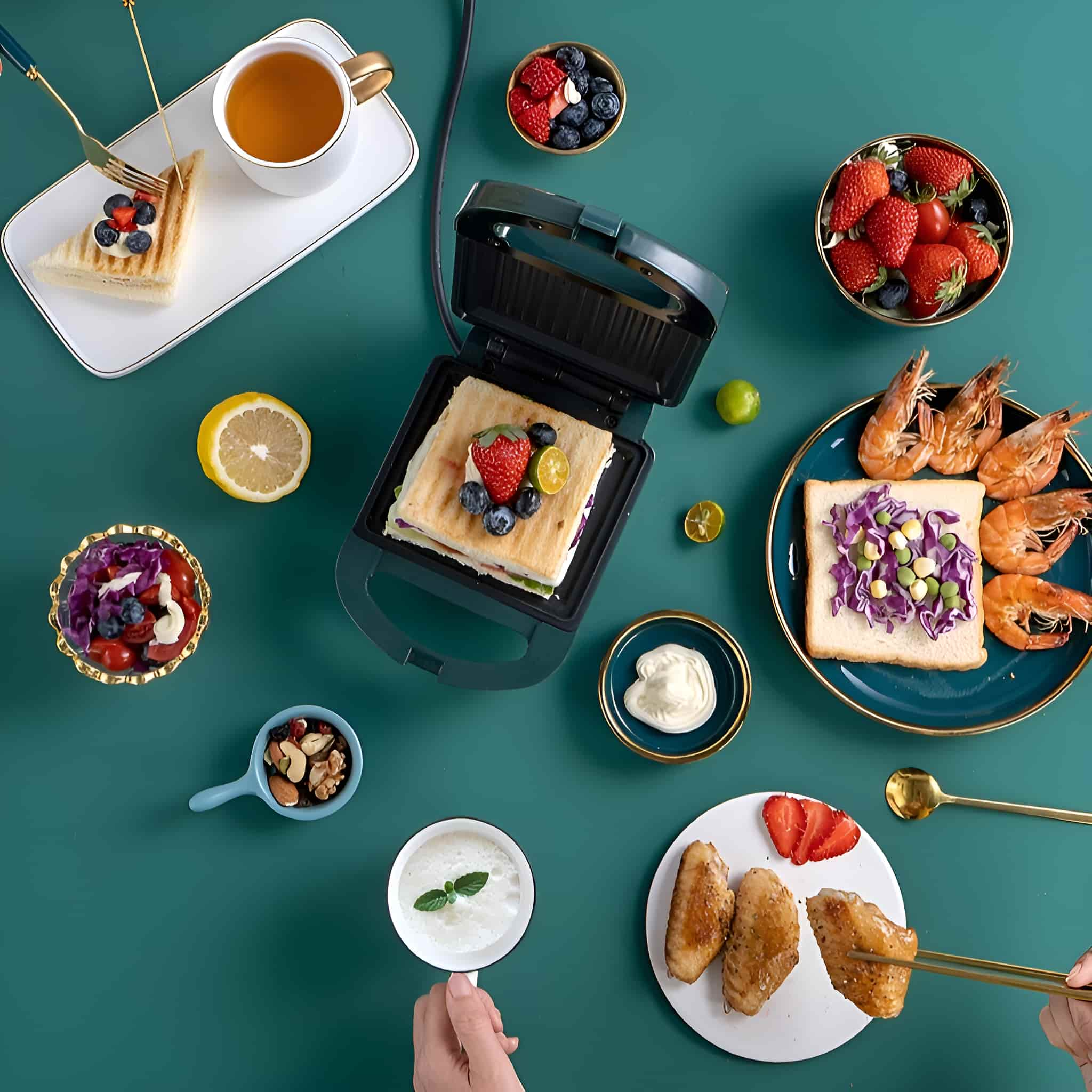 Tisch mit verschiedenen Speisen: Sandwich, Erdbeeren, Garnelen, Flügel, Nüsse und Joghurt. Ein Sandwich liegt auf einem Sandwichmaker.
