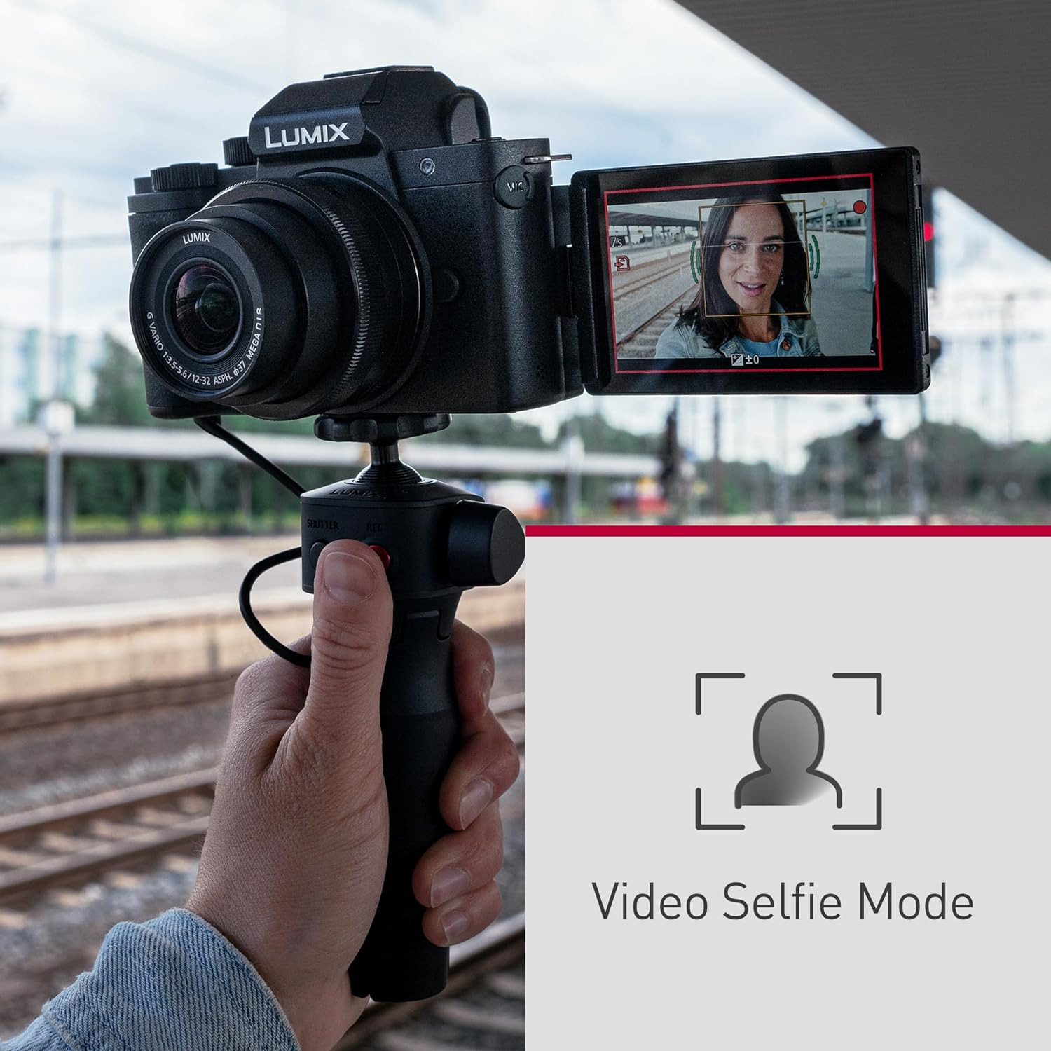 Eine Person hält eine Kamera mit geöffnetem Selfie-Bildschirm. Der Hintergrund ist ein Bahnhof.