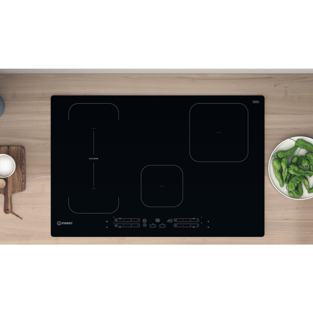Czarna płyta indukcyjna z czterema strefami gotowania. Zielone papryki na drewnianej desce.