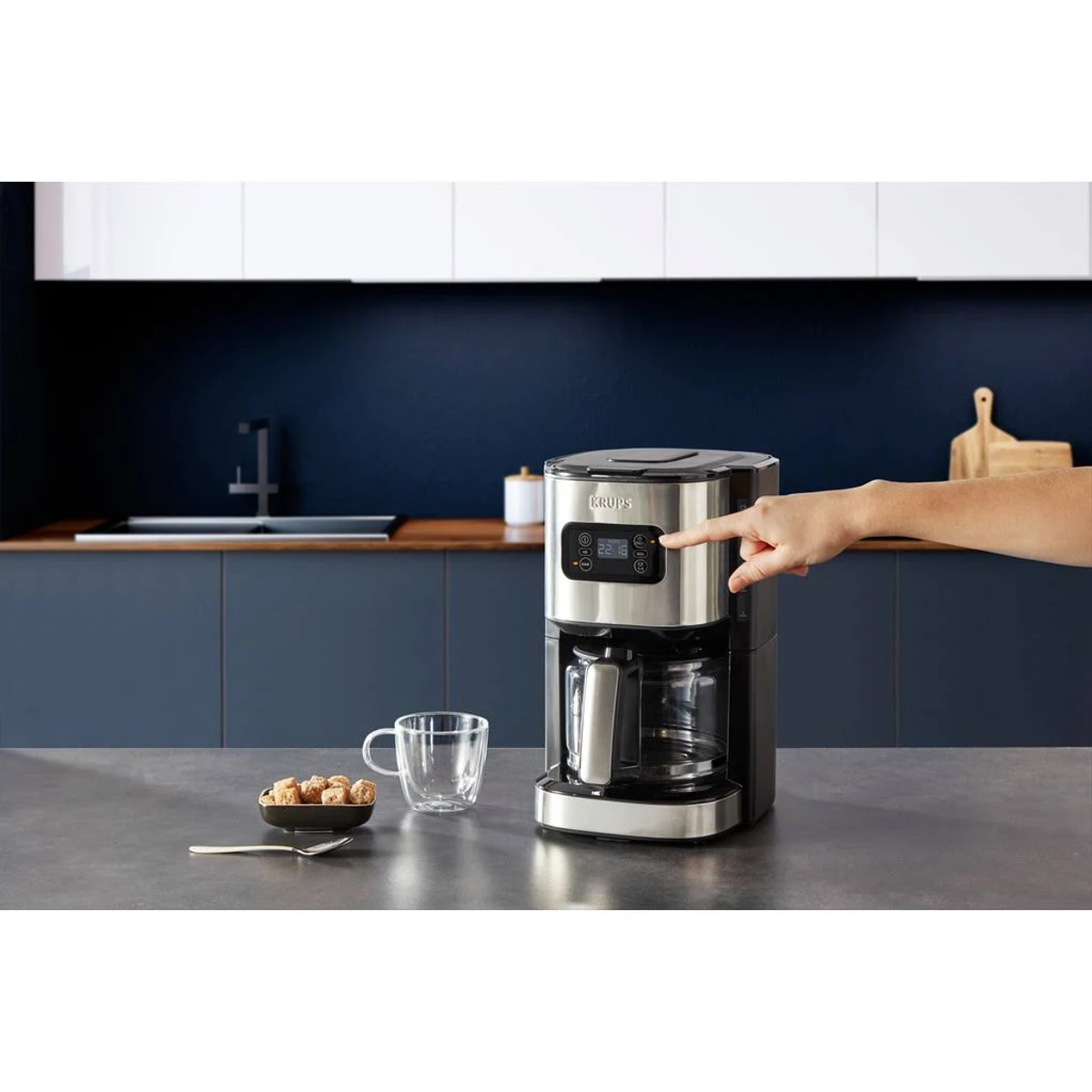 Macchina da caffè con la mano di una persona che preme un pulsante. Caffè, tazza e cucchiaio visibili.