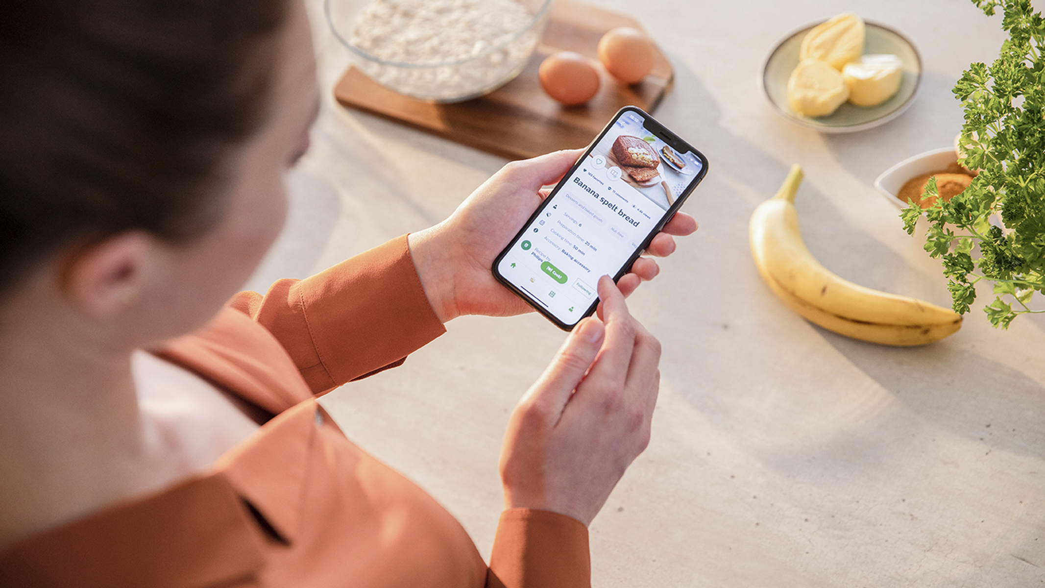 Eine Person hält ein Telefon, das ein Rezept für Bananenbrot zeigt. Zutaten im Hintergrund.