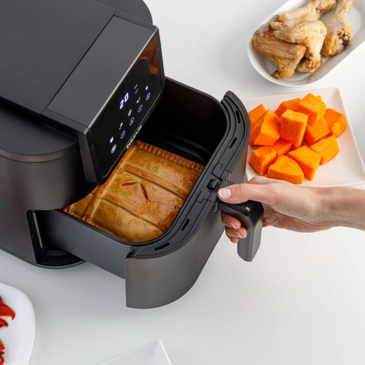 Czarna frytkownica beztłuszczowa z jedzeniem w środku i dynią oraz skrzydełkami obok na białym stole.